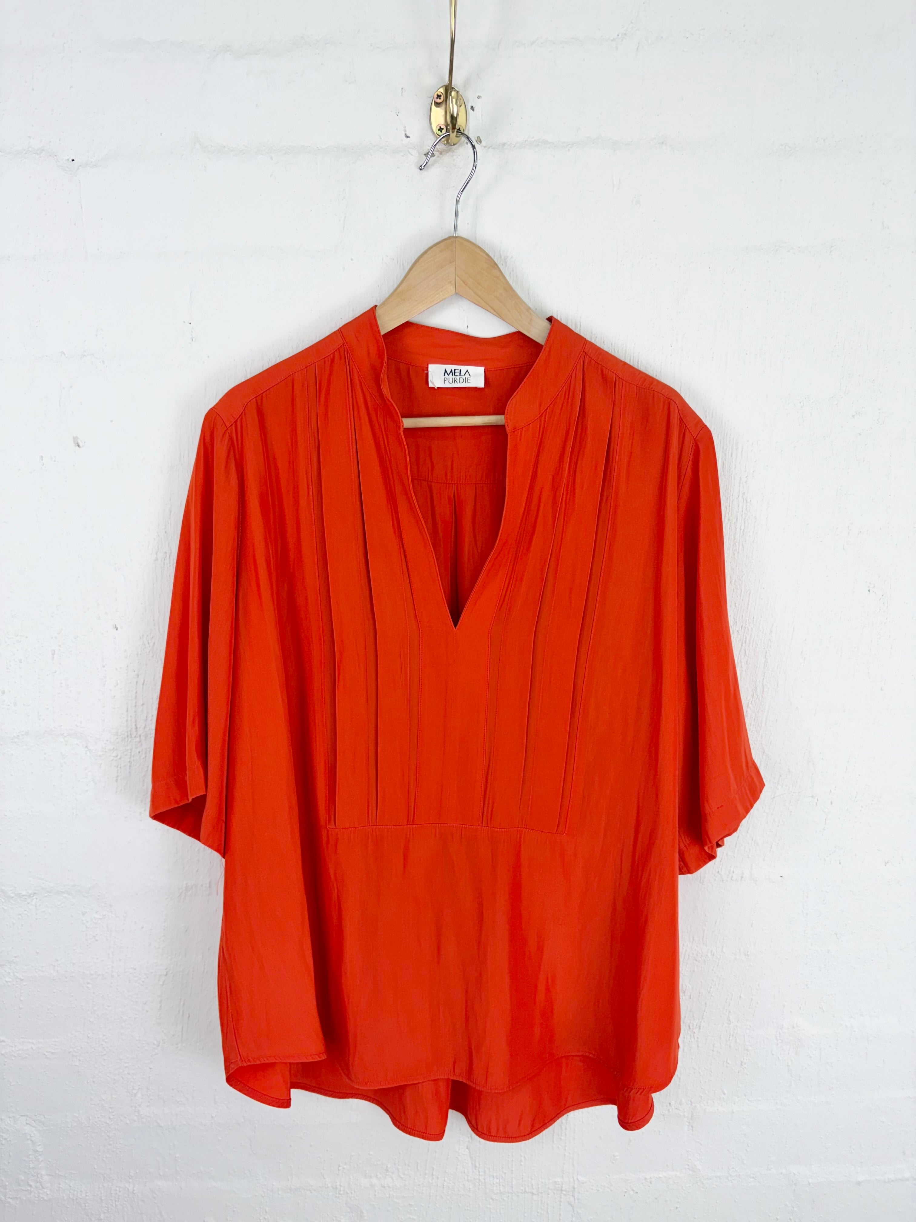 A bright orange-red short-sleeve blouse with a V-neckline and pleated front, hanging on a wooden hanger against a white painted brick wall.