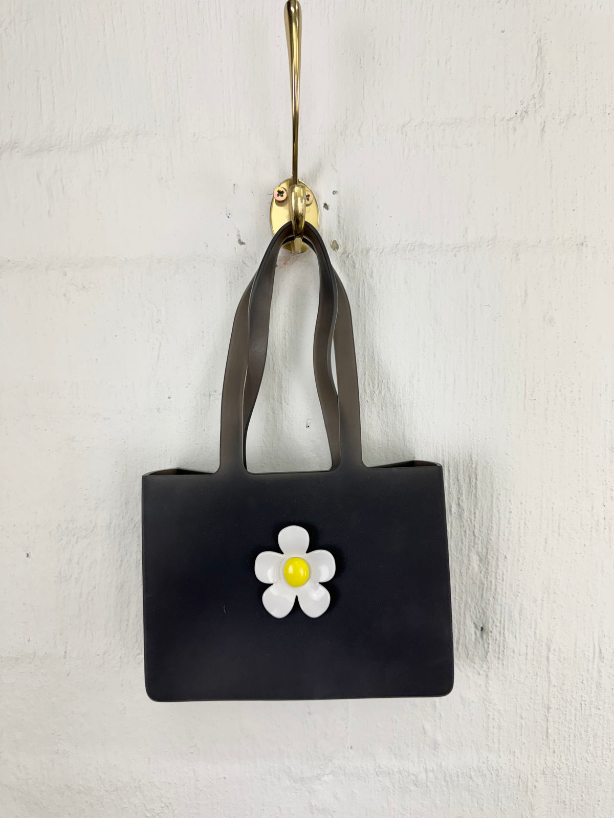 A small black tote bag with dark gray handles and a white flower applique with a yellow center, hanging from a gold hook on a white textured wall.