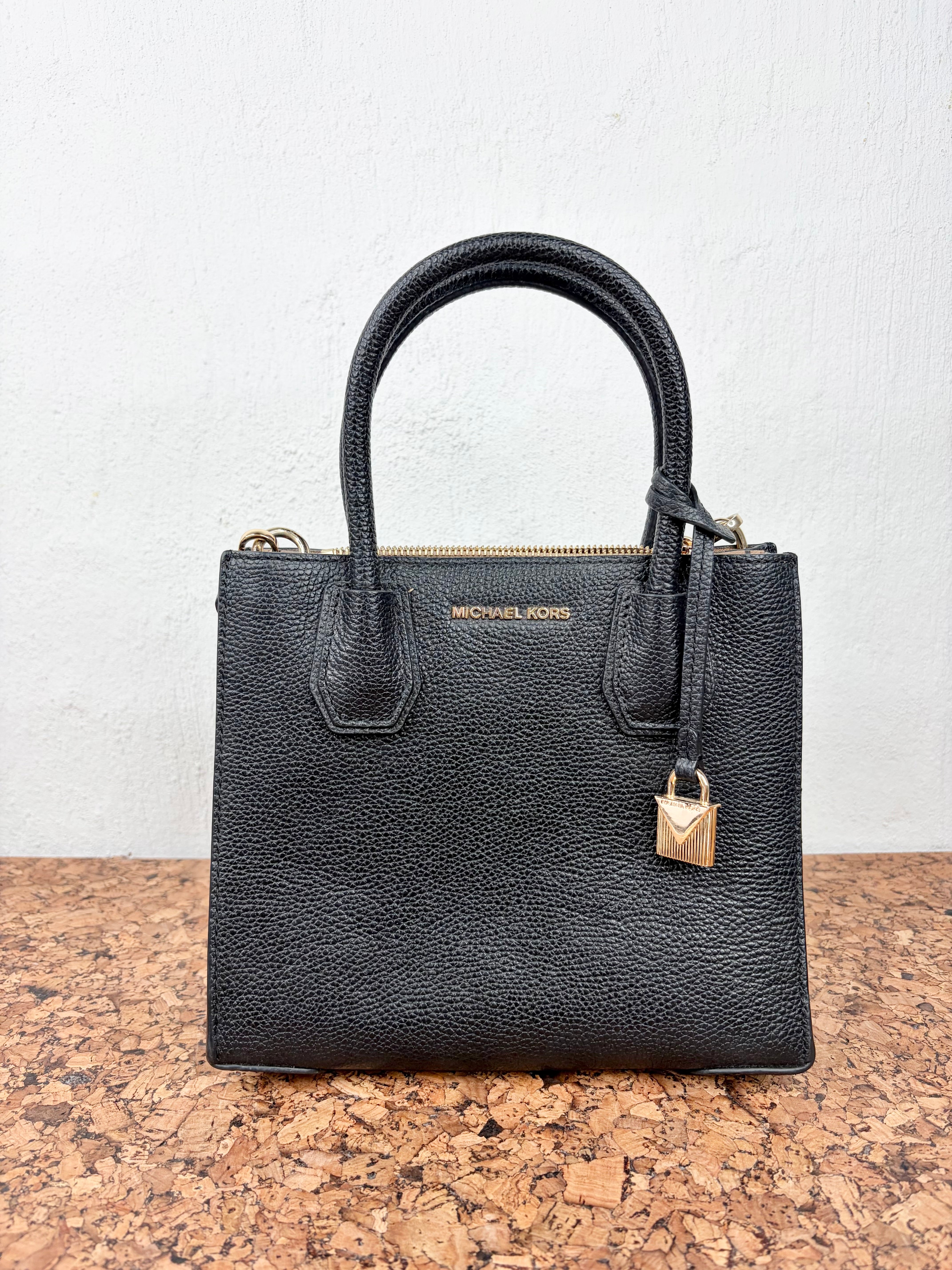 A black pebbled leather handbag by Michael Kors with dual top handles, a top zipper, and a gold padlock charm, displayed on a cork surface against a white wall.