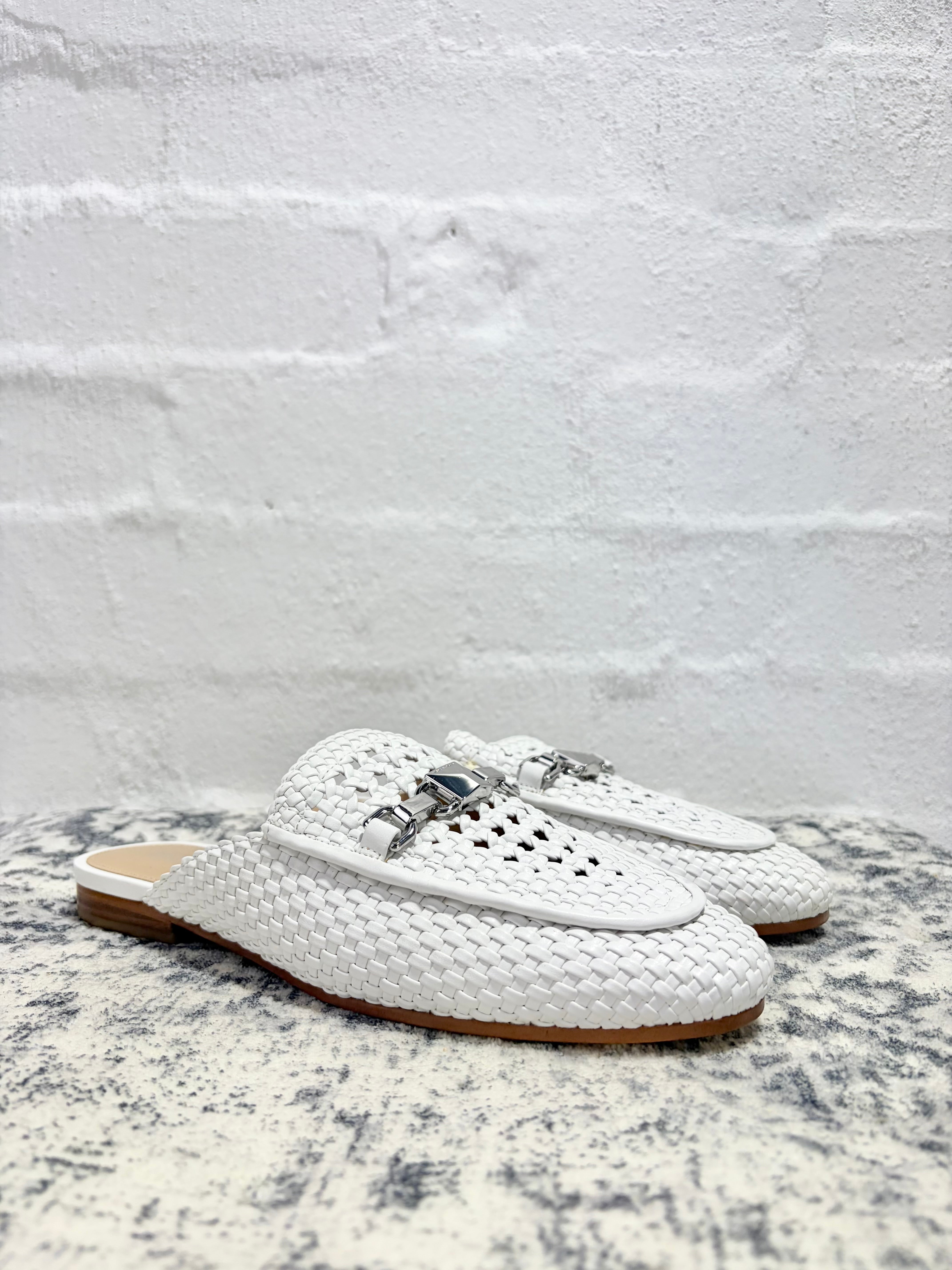 A pair of white woven leather mule loafers with silver-tone metal hardware, displayed on a textured gray surface against a white brick wall background.