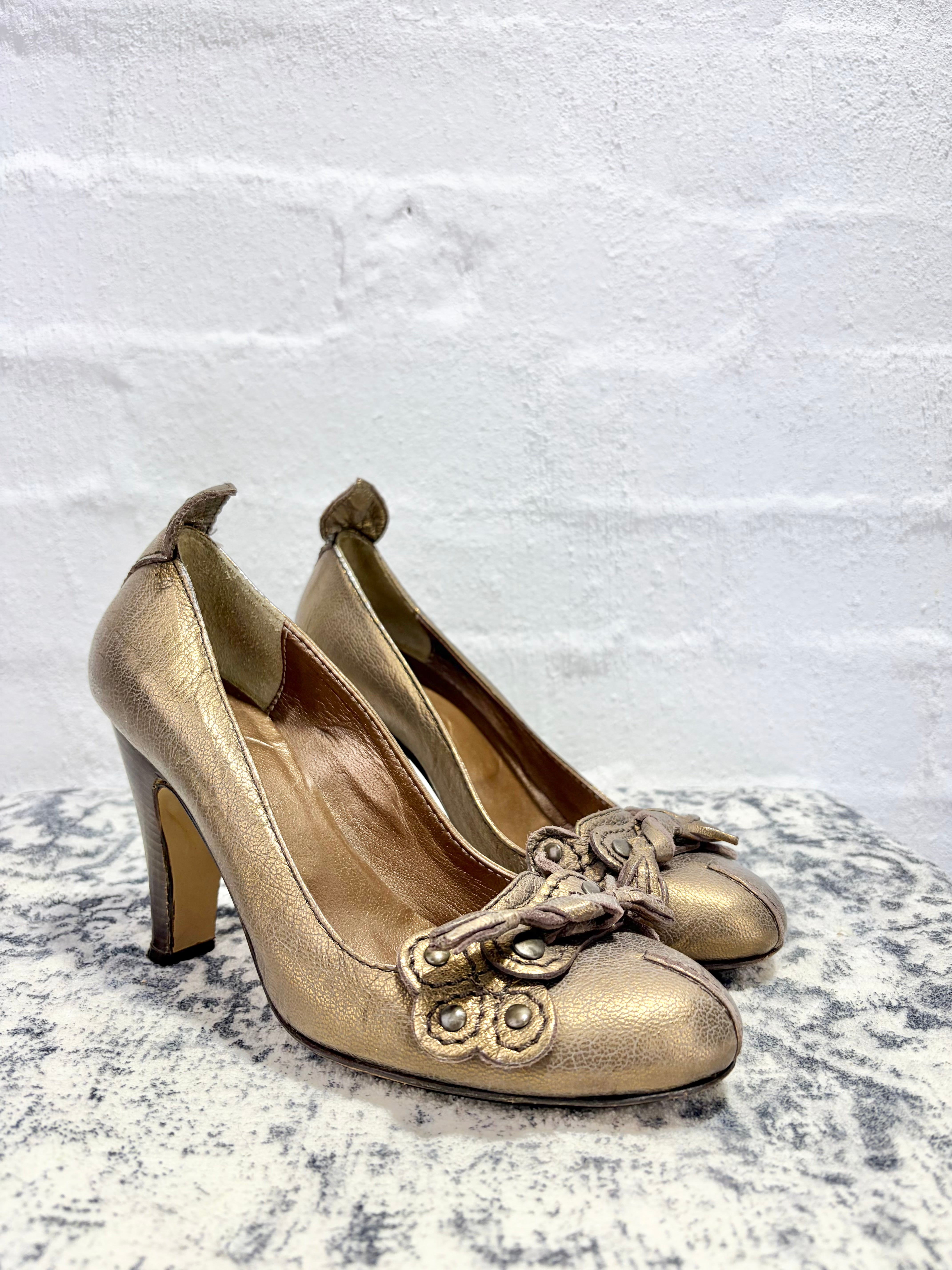 A pair of metallic gold high-heeled pumps with ornate baroque-style metallic embellishments on the toe, displayed on a textured gray surface against a white brick wall.