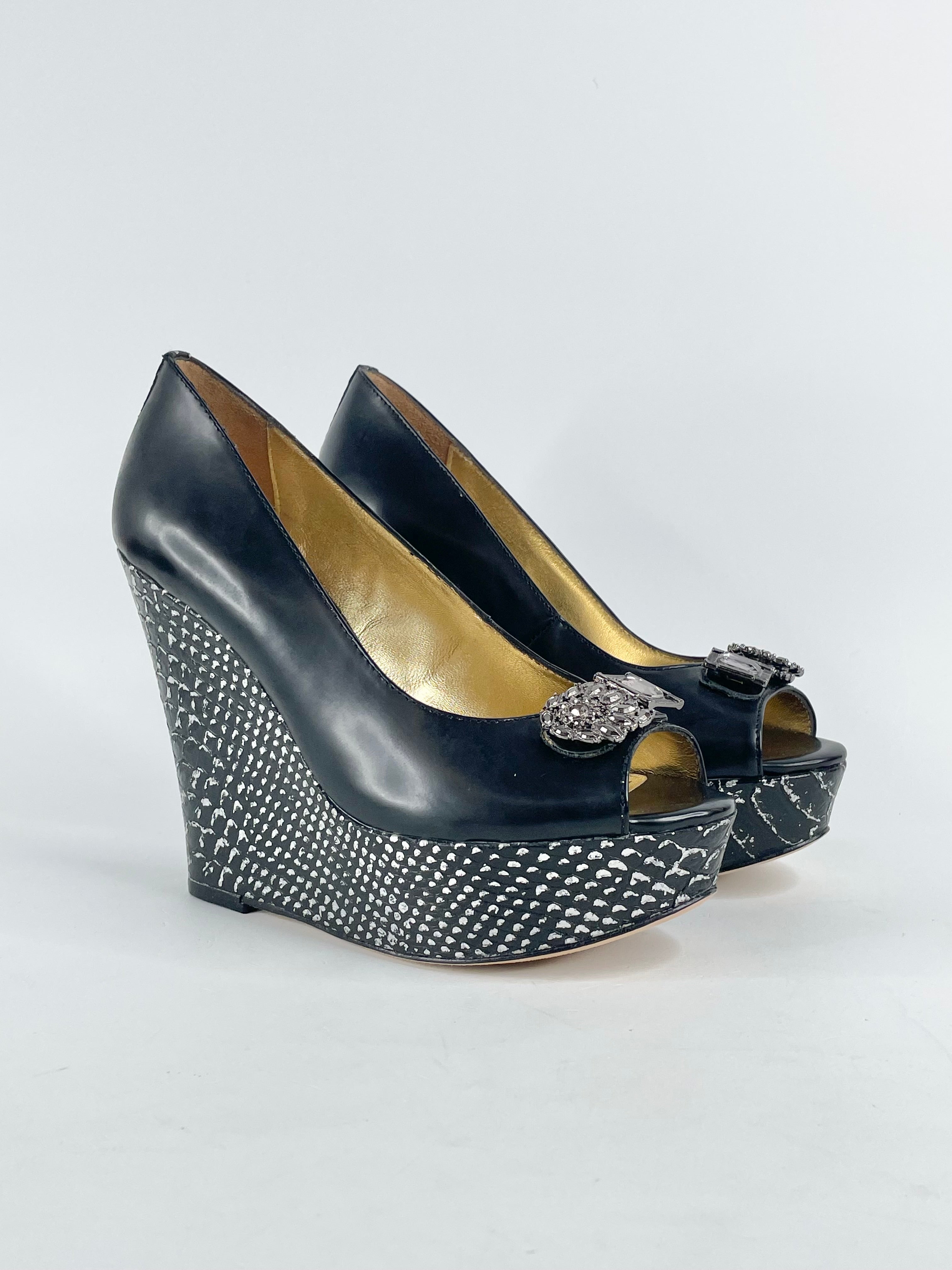 A black leather wedge shoe with golden contrasts, featuring a decorative brooch on the toe and a textured pattern.