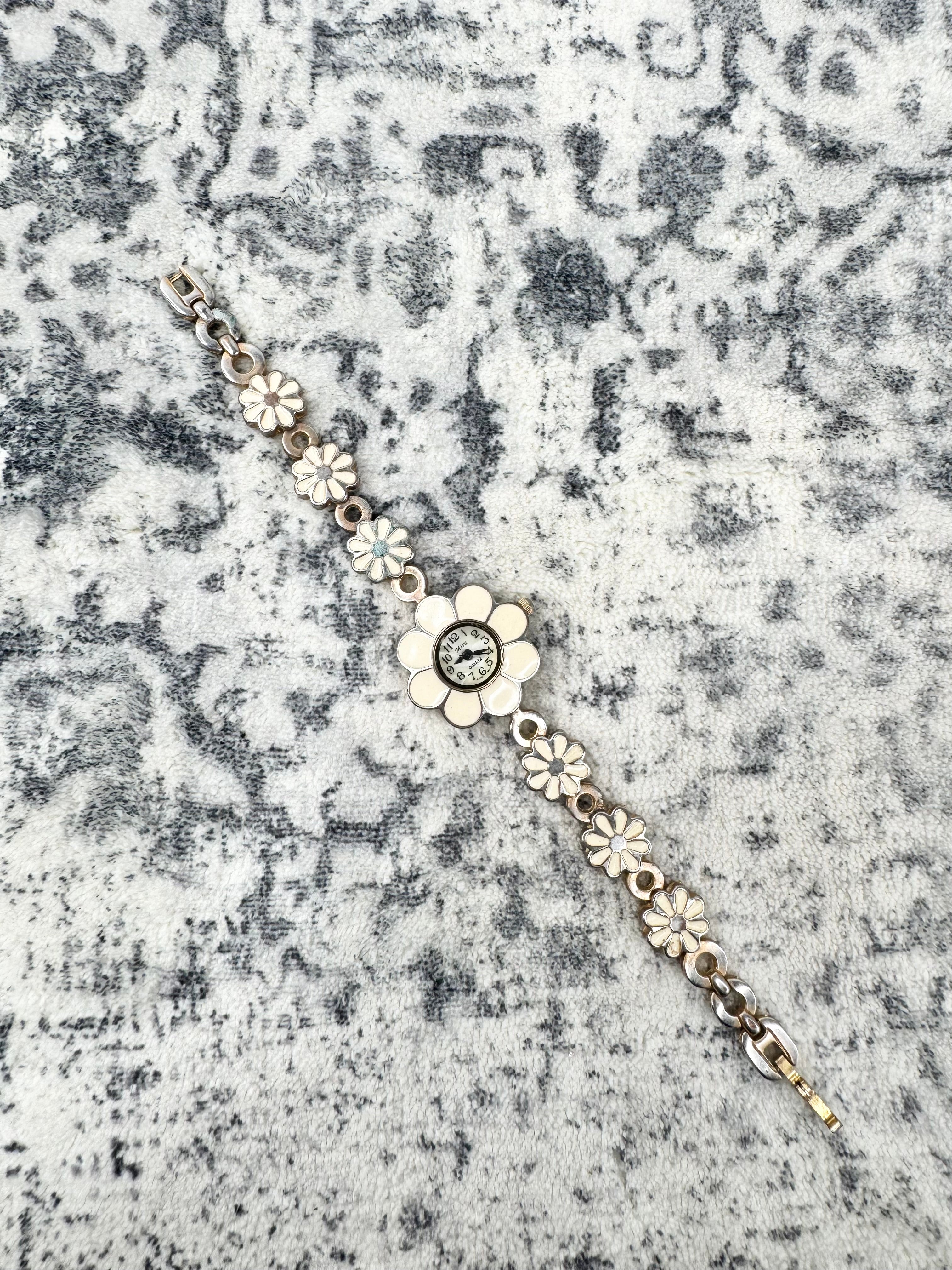 A silver-tone floral bracelet featuring a central flower with a clear stone and smaller flower motifs along the chain, displayed on a textured gray and white surface.