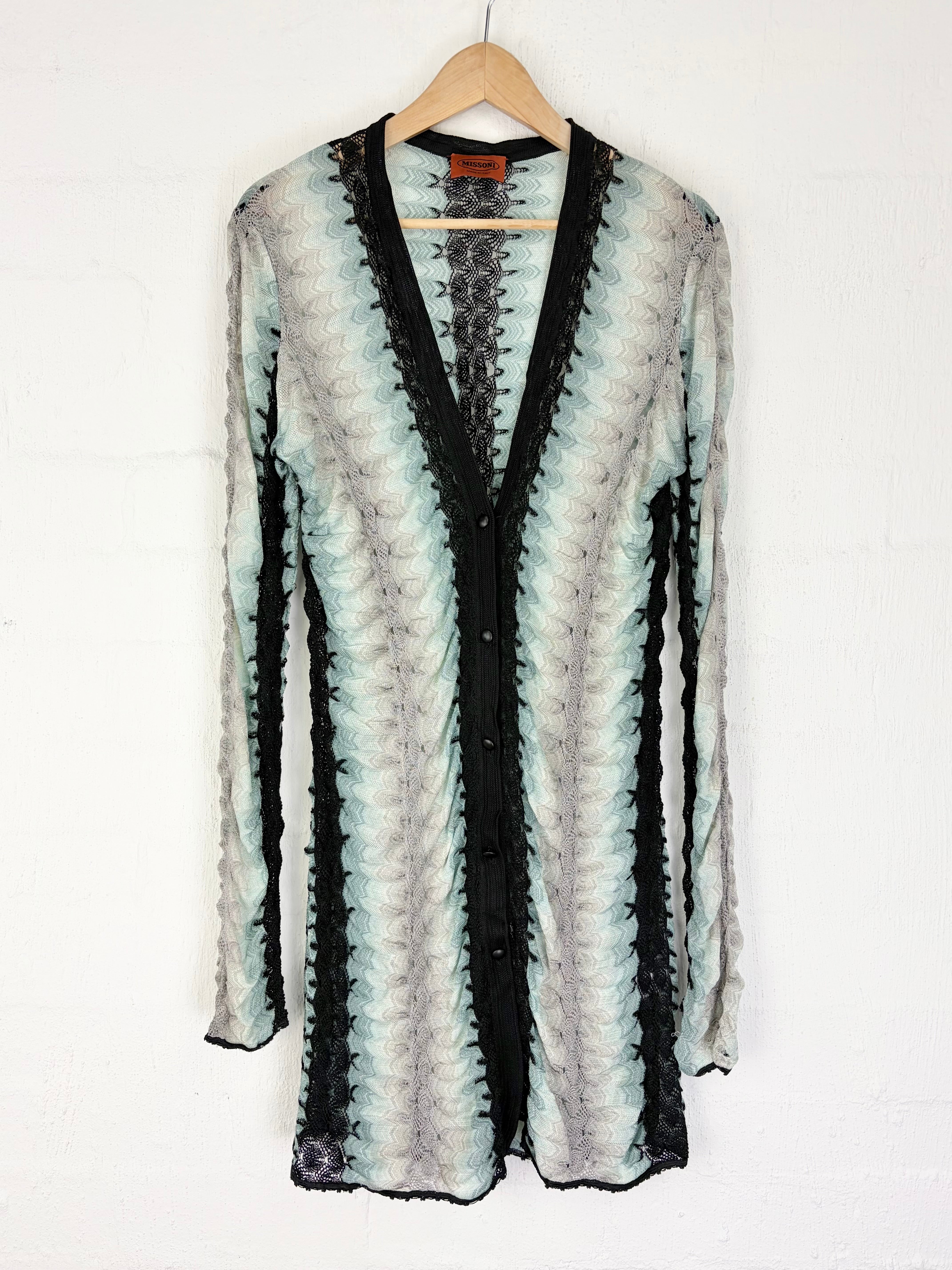 A long-sleeve open-front cardigan hanging on a wooden hanger against a white wall. The cardigan features a vertical wavy pattern in black, light gray, and pale mint green, with solid black lace trim along the V-neckline and button placket.
