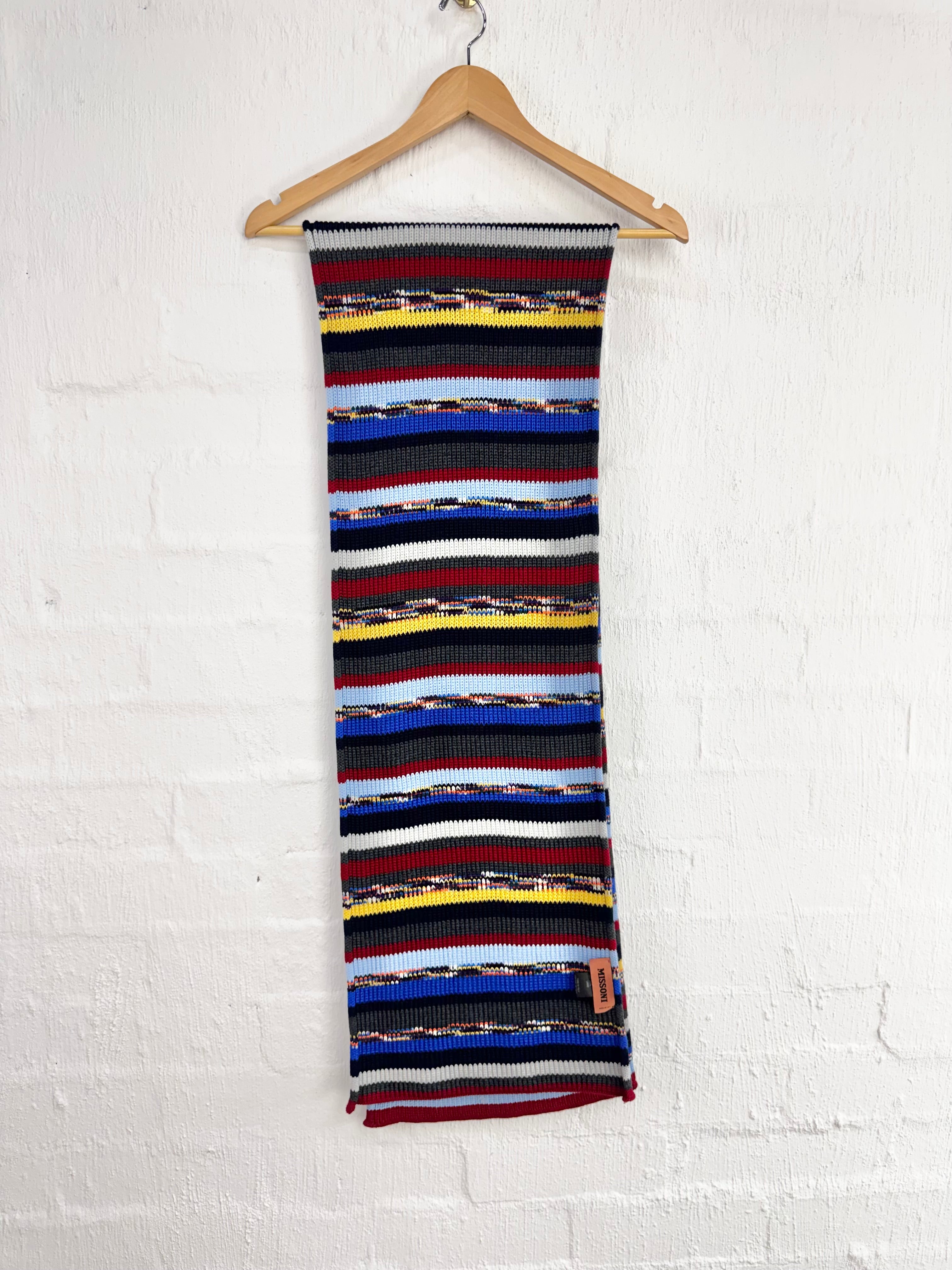 A long knit scarf with horizontal multicolor stripes, hanging from a wooden hanger against a white painted brick wall.