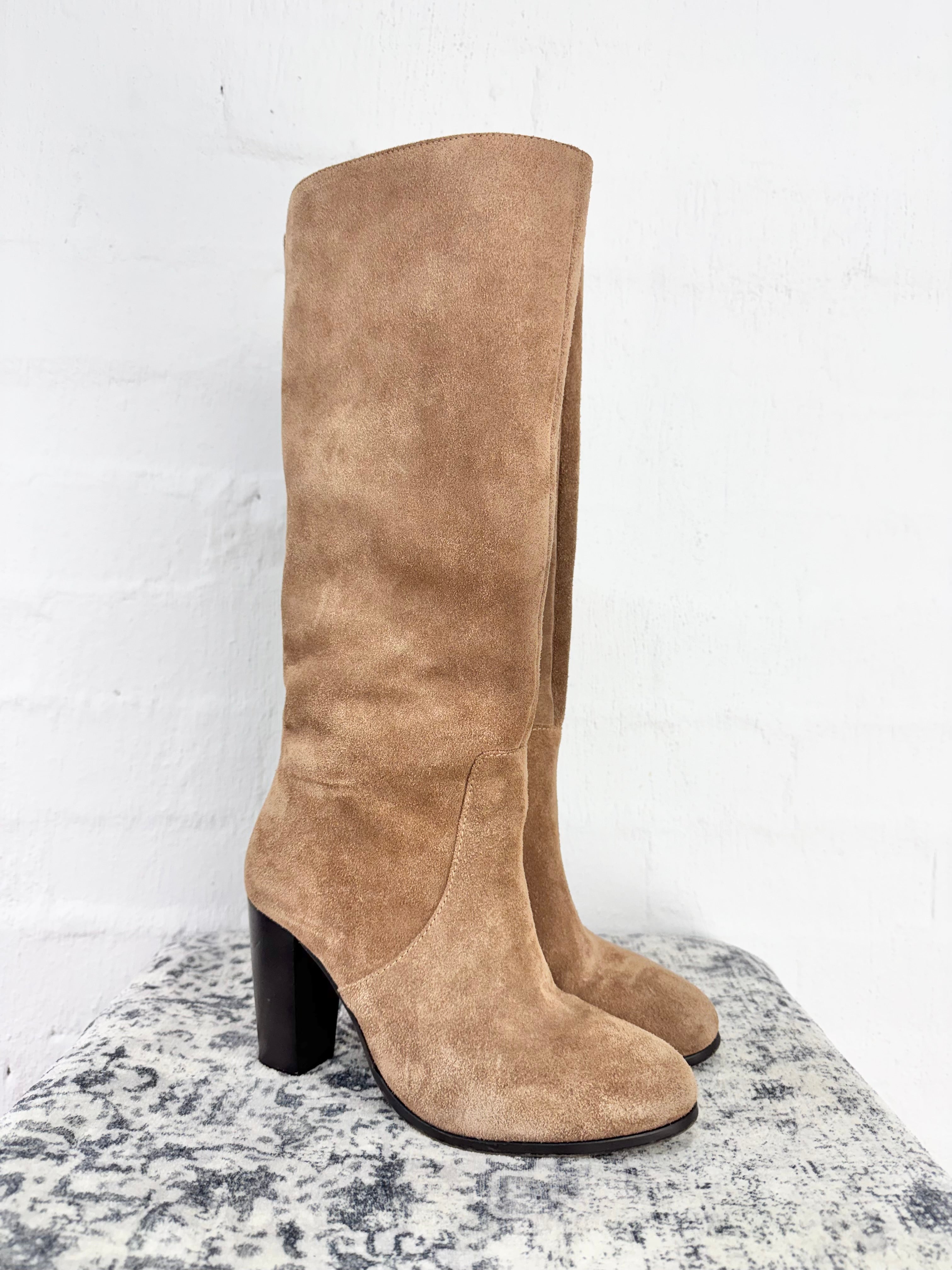 A pair of tall knee-high suede boots in a light taupe-brown color, featuring a chunky black block heel, displayed on a textured gray surface against a white brick wall.