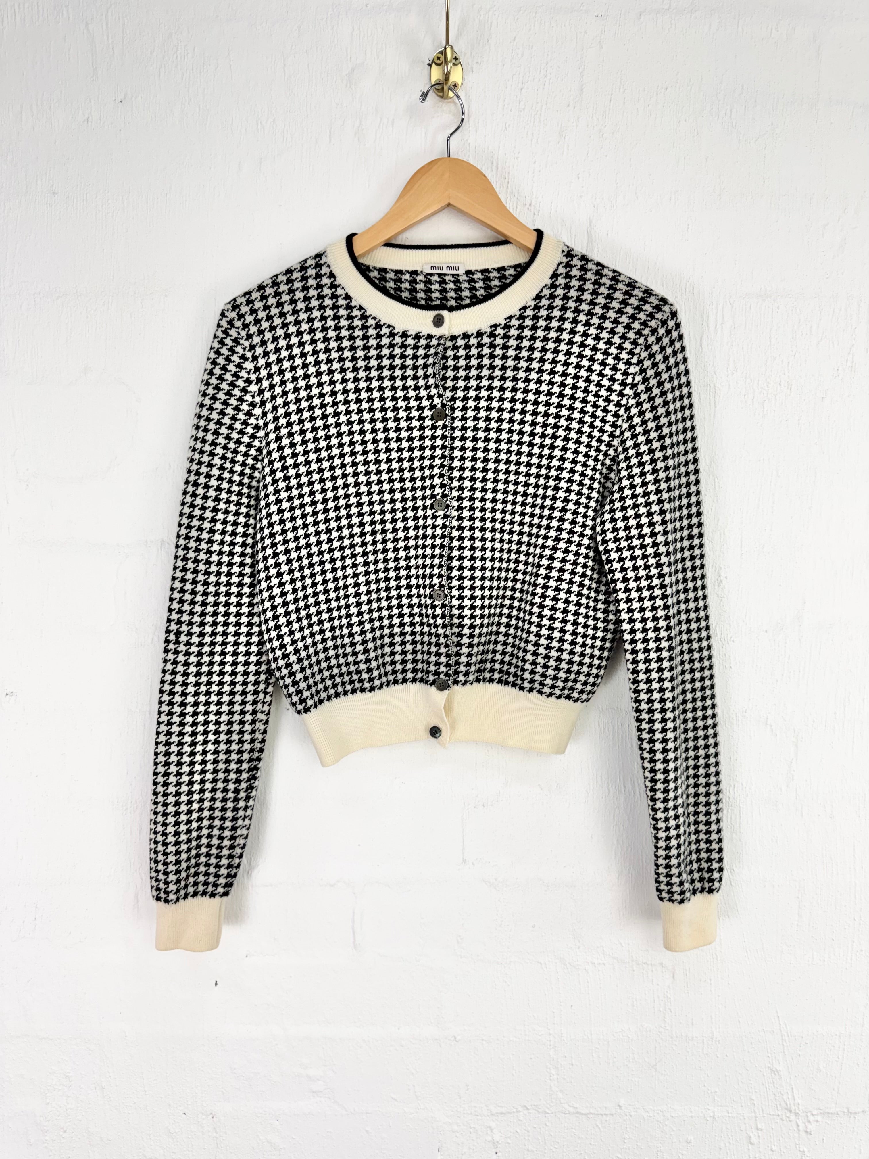 A cropped long-sleeve cardigan with a black and white houndstooth pattern, cream ribbed collar, hem, and cuffs, hanging on a wooden hanger against a white brick wall.