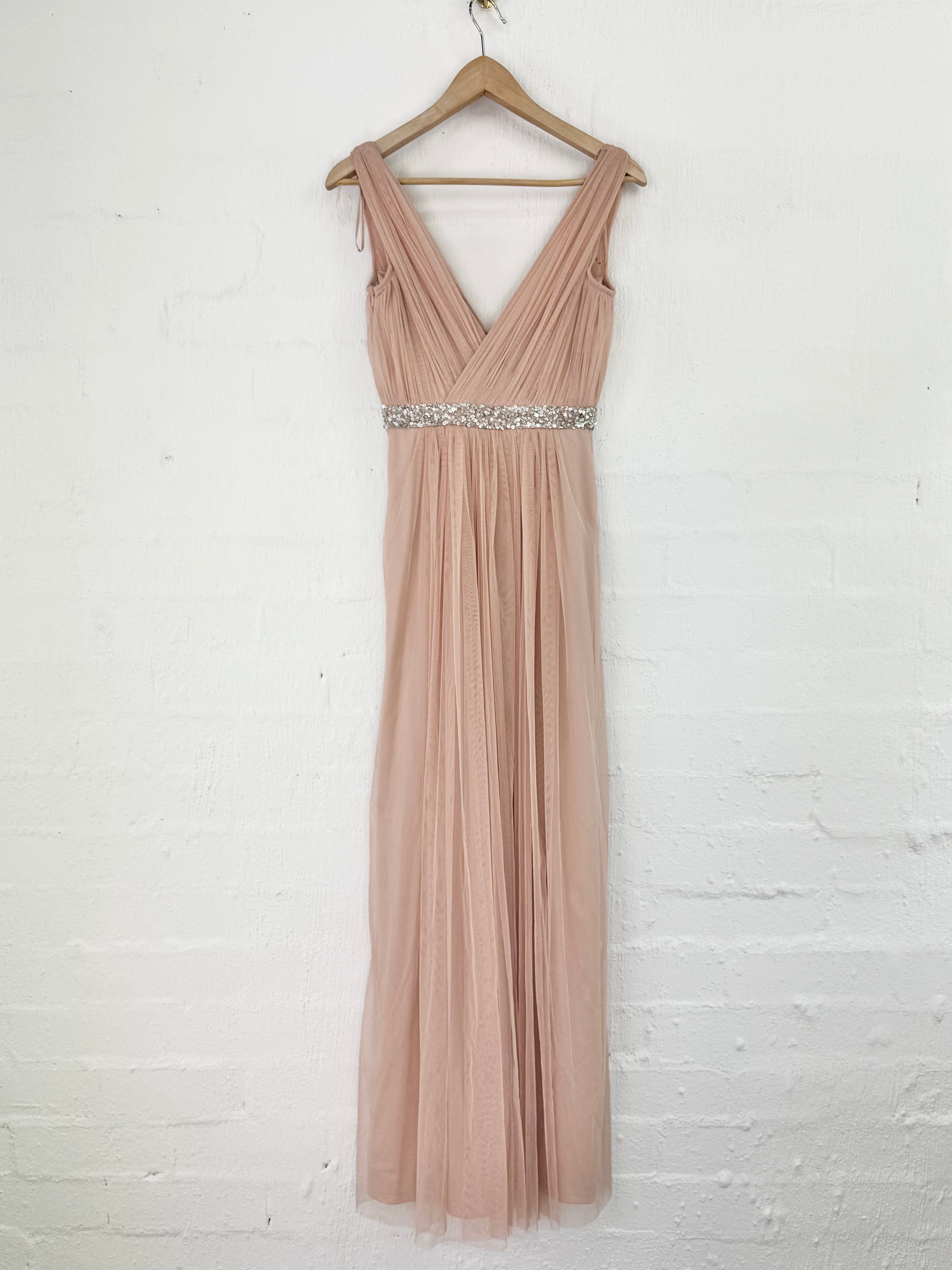 A sleeveless, floor-length blush pink tulle gown with a V-neckline and a sparkling silver beaded waistband, hanging on a wooden hanger against a white painted brick wall.