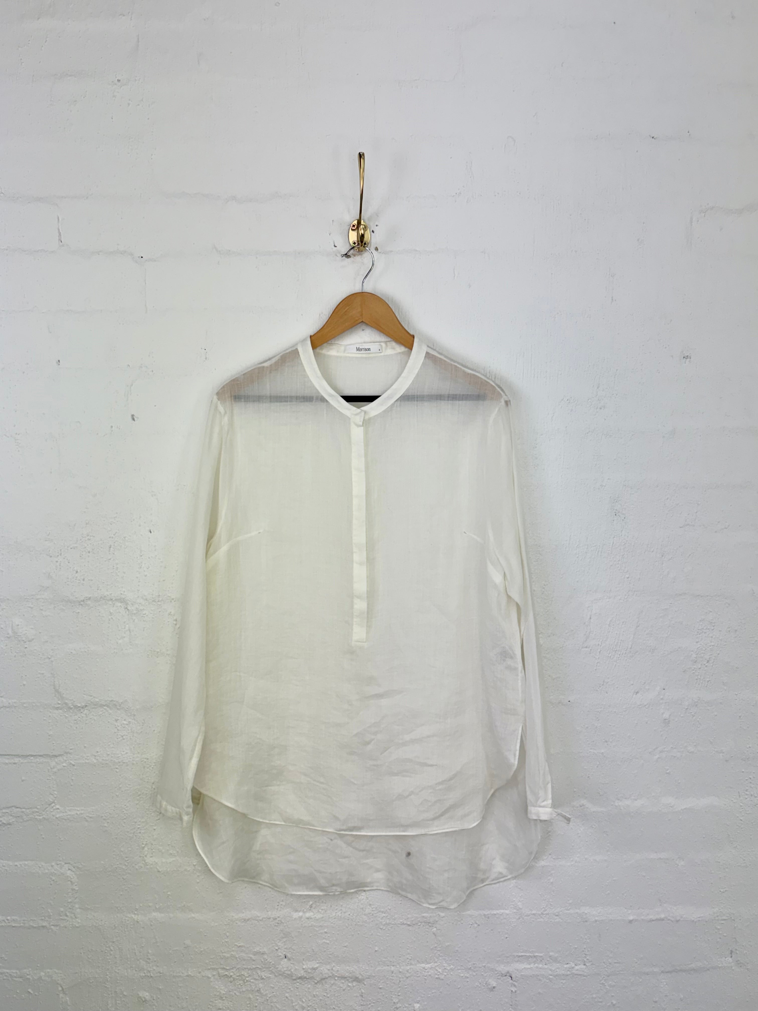 A long-sleeve, semi-sheer white blouse with a mock neck and button placket, hanging on a wooden hanger from a gold hook against a white brick wall.