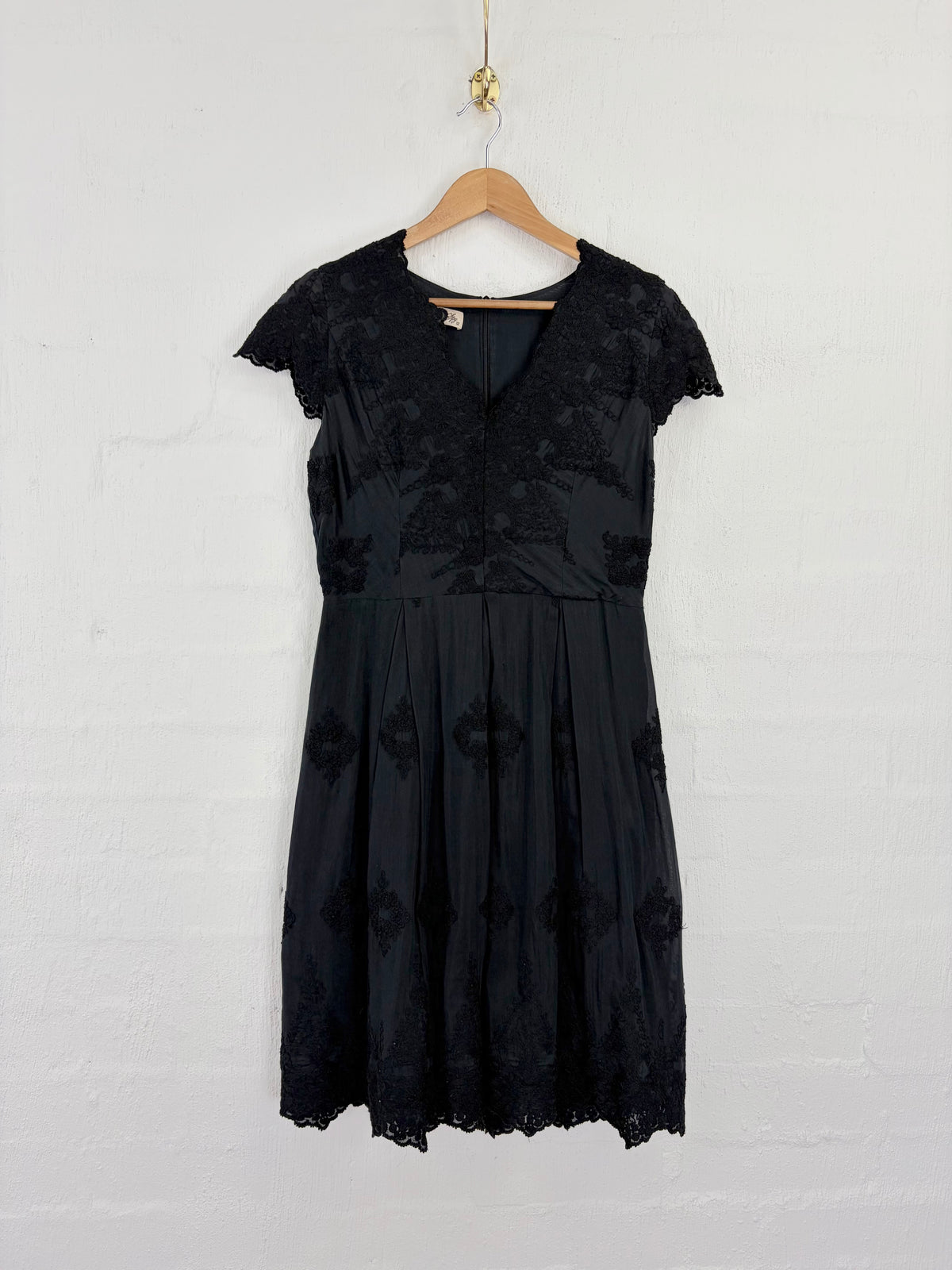 A black lace and mesh A-line dress with short cap sleeves, hanging on a wooden hanger against a white painted brick wall.