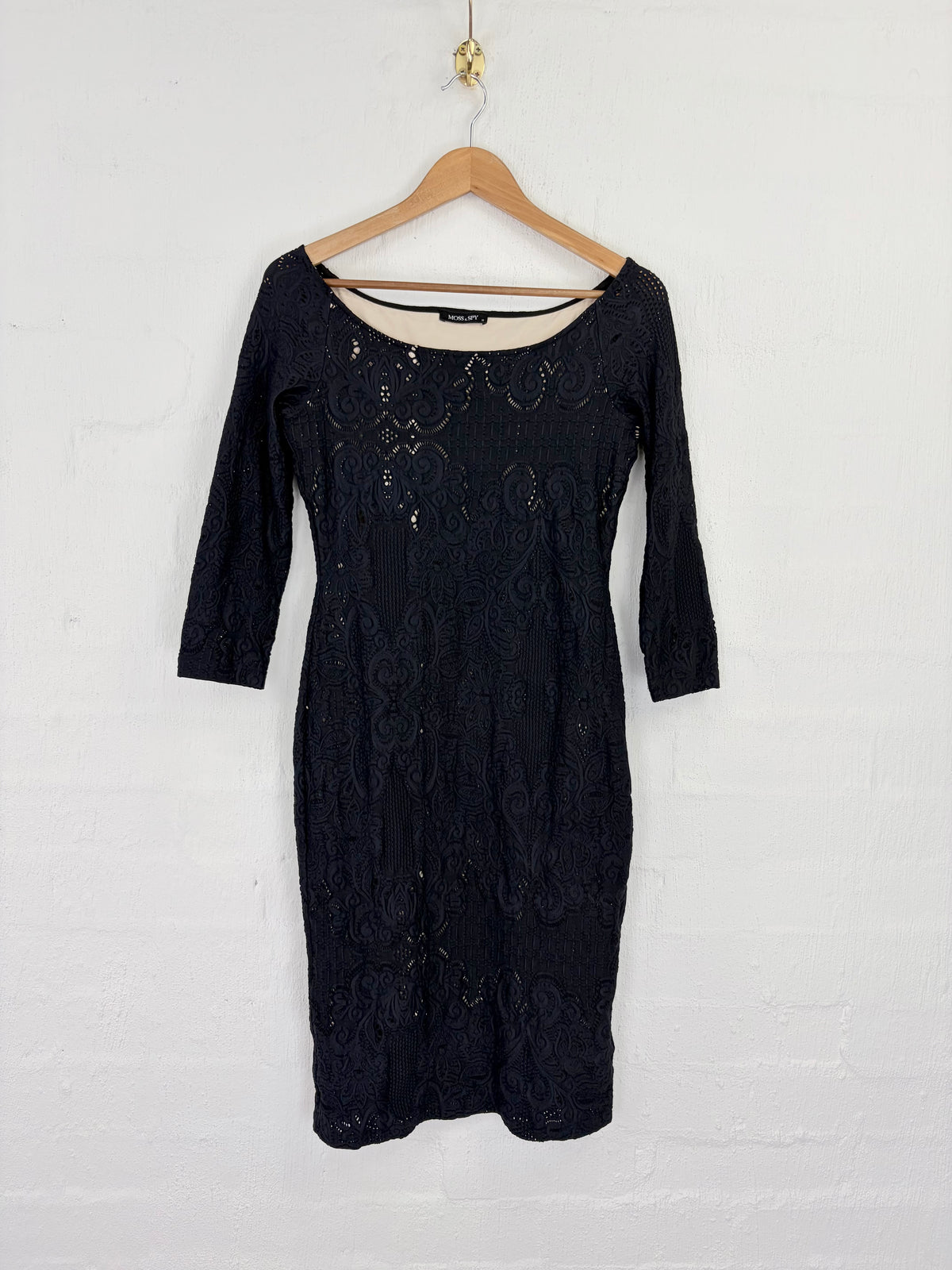 A black lace three-quarter sleeve dress hanging on a wooden hanger against a white painted brick wall.