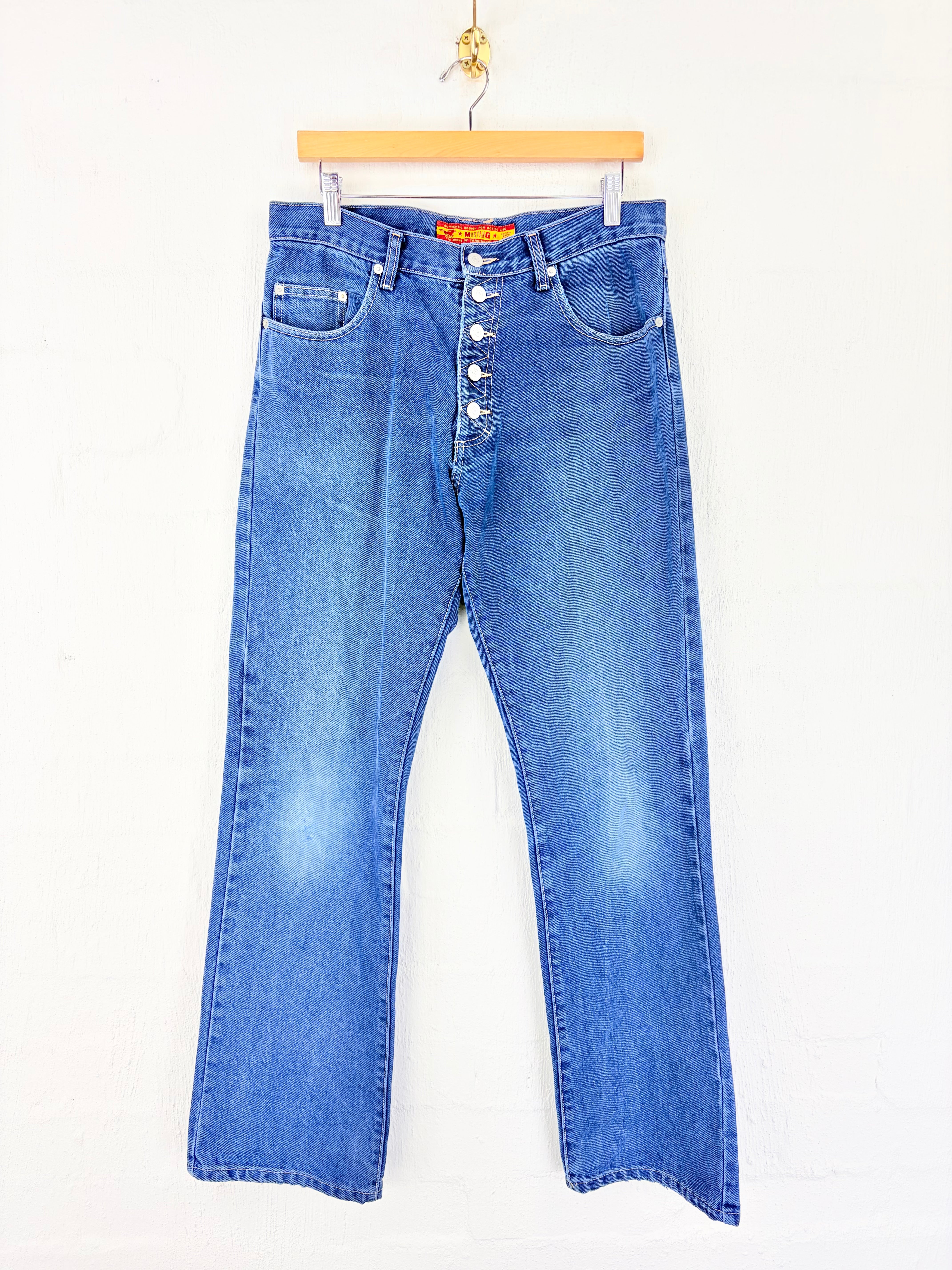 A pair of blue denim jeans with a button fly, hanging from a wooden hanger against a white wall.