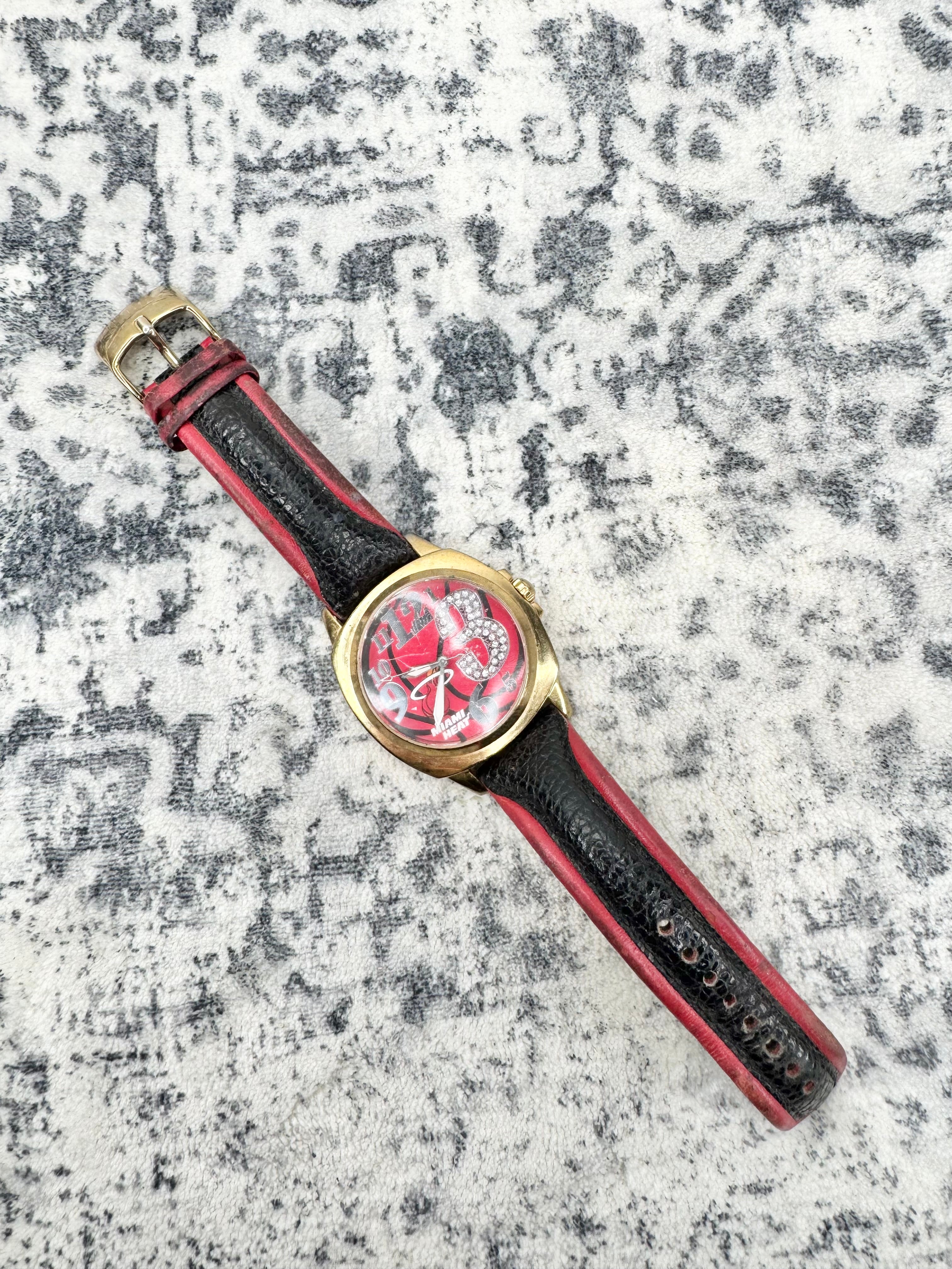 A wristwatch with a gold-tone case, a red and white patterned dial, and a black leather strap with red stitching, displayed diagonally on a textured gray and white surface.