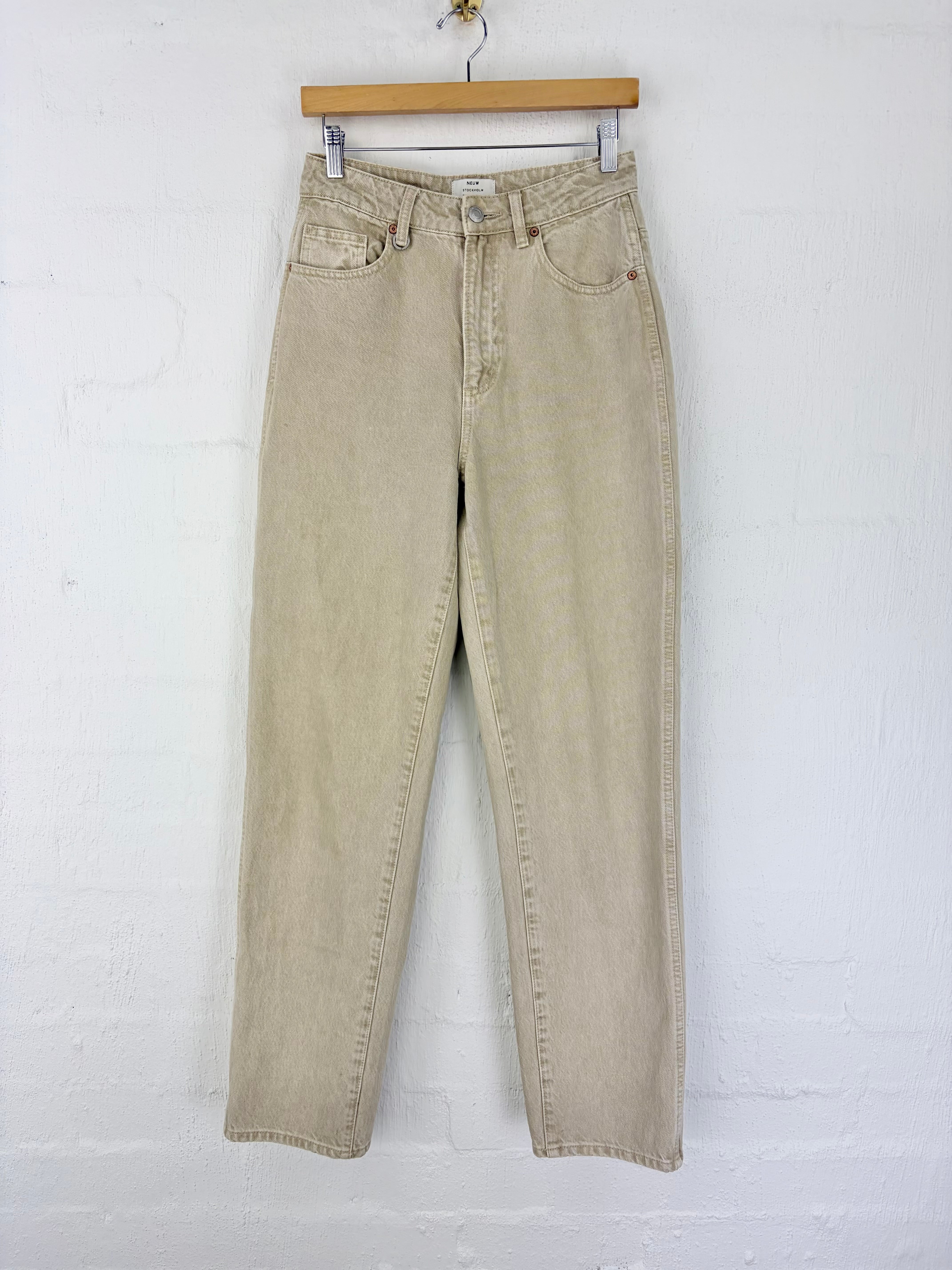 A pair of light beige denim jeans hanging from a wooden hanger against a white painted brick wall.