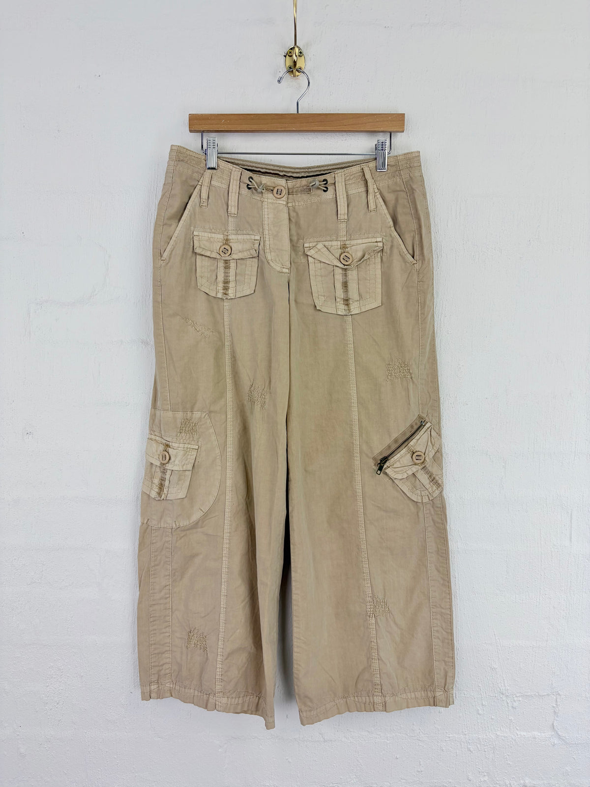 A pair of beige corduroy cargo pants with multiple flap pockets, hanging from a wooden hanger against a white painted brick wall.
