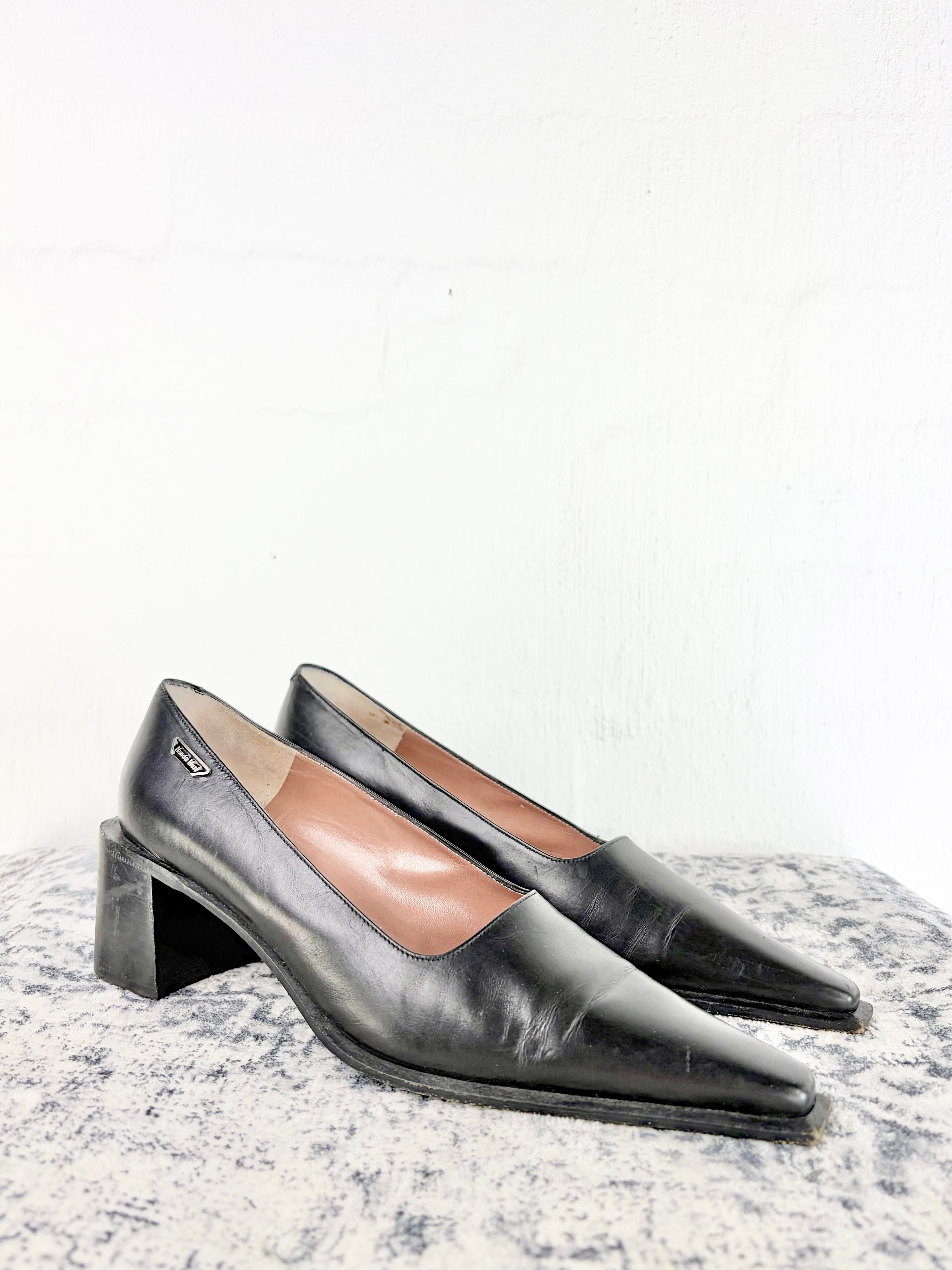 A pair of black leather pointed-toe pumps with block heels, displayed on a textured gray surface against a white wall.