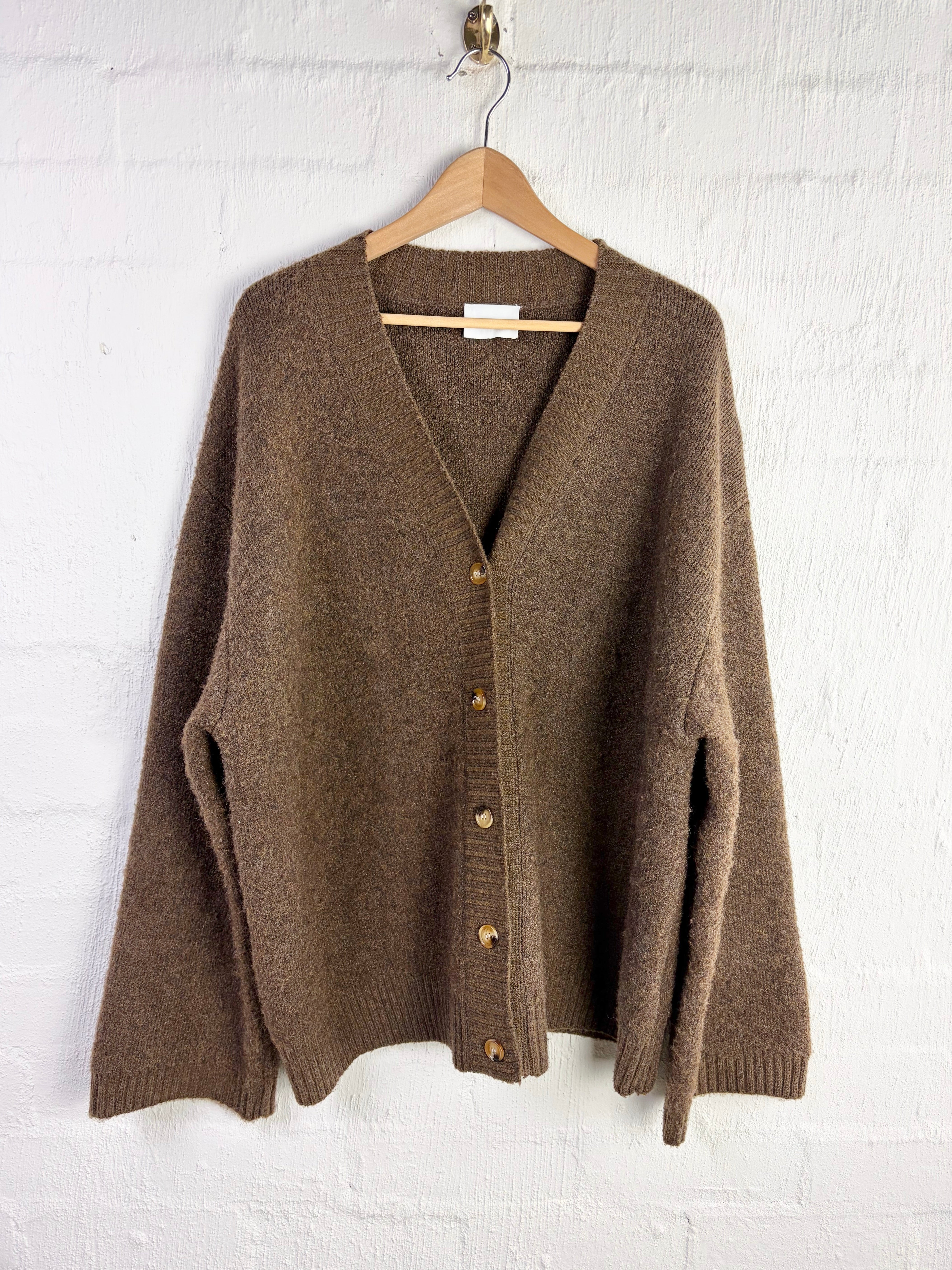 A taupe brown knit cardigan sweater with a V-neck and button-front closure, hanging on a wooden hanger against a white painted brick wall.