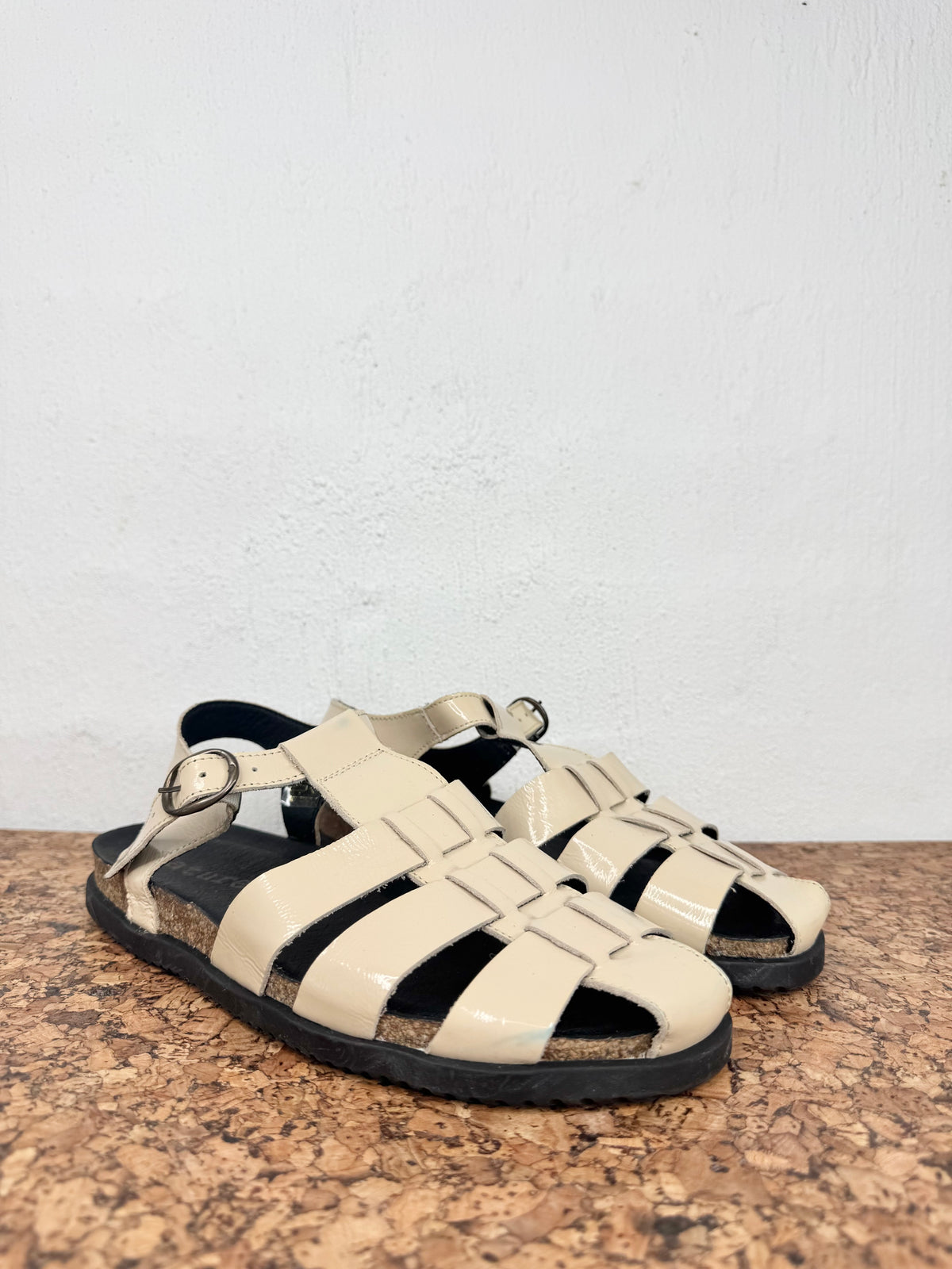 A pair of beige woven-look strappy sandals with black soles, displayed on a cork surface against a white wall.