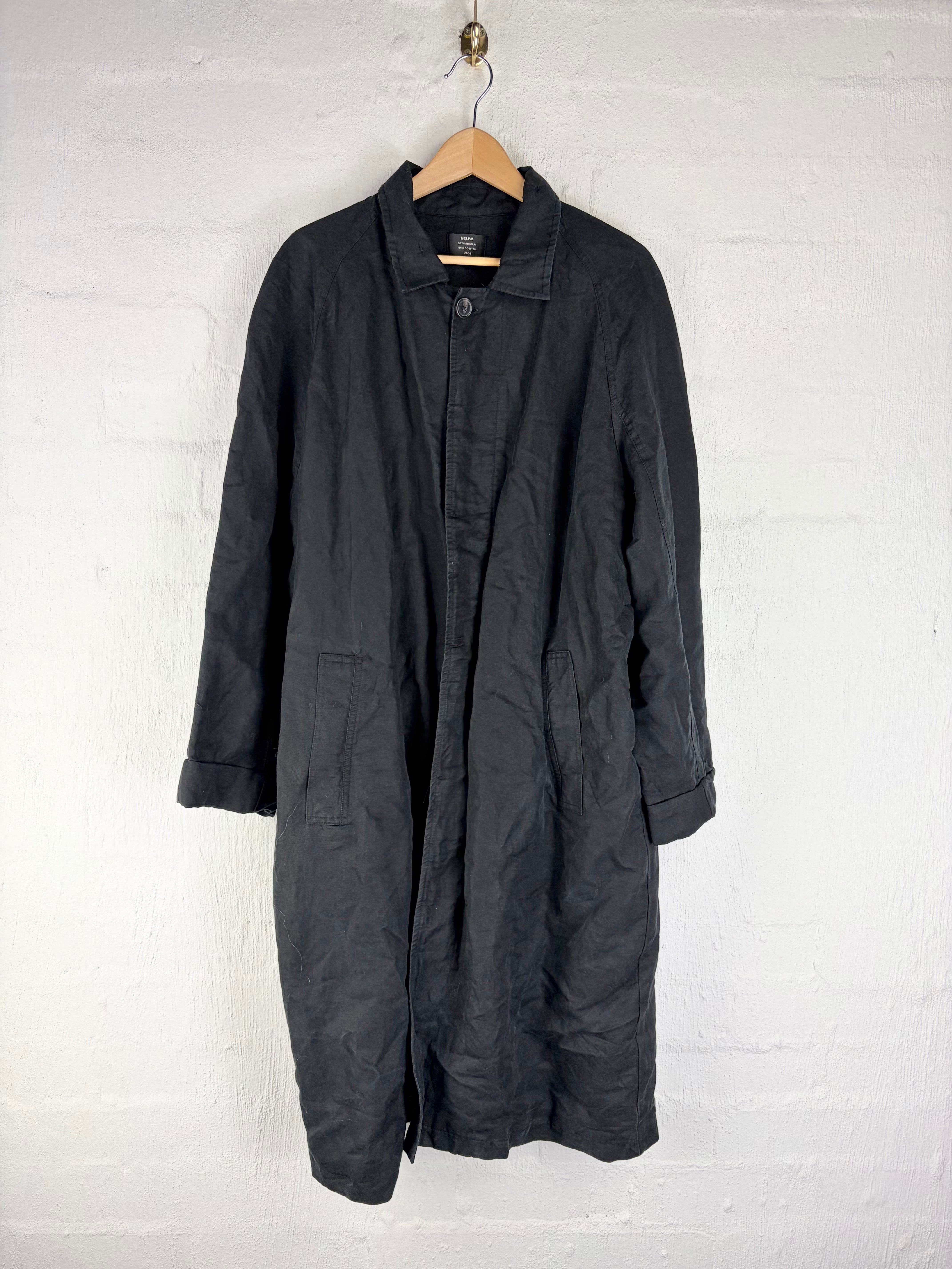 A black mid-length coat with a relaxed, slightly wrinkled texture, hanging on a wooden hanger against a white painted brick wall.