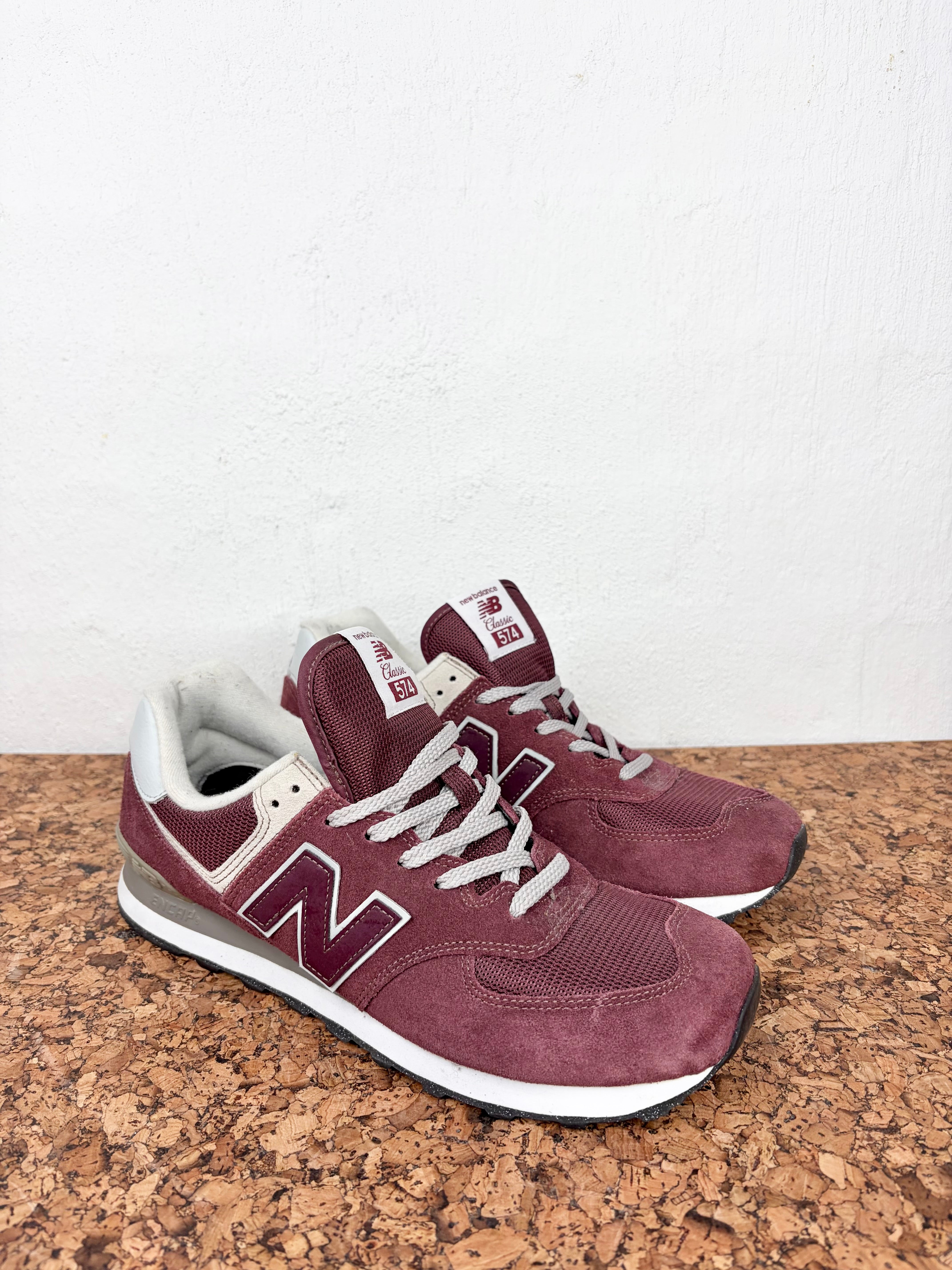 A pair of New Balance 574 sneakers in burgundy suede and mesh with white laces and a white midsole, resting on a cork mat against a white wall.
