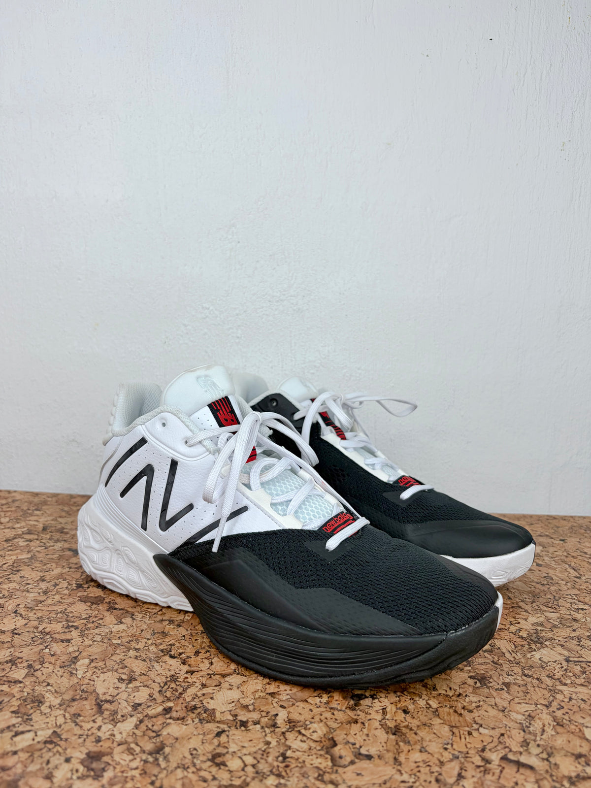 A pair of New Balance 1000 sneakers in black, white, and red, displayed on a textured brown surface against a plain white wall.