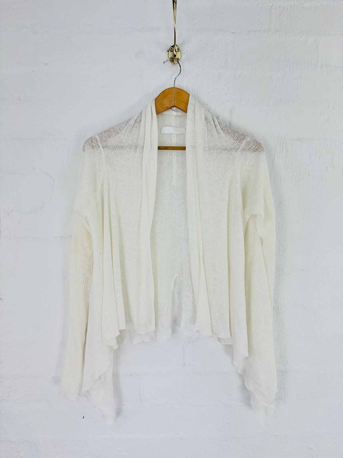 An off-white open-front cardigan sweater with a fuzzy knit texture and long draped sleeves, hanging on a wooden hanger against a white painted brick wall.