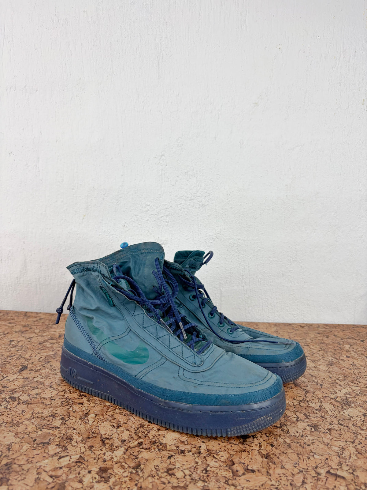 A pair of teal-blue high-top sneakers with matching blue laces and soles, resting on a brown speckled surface against a white wall.