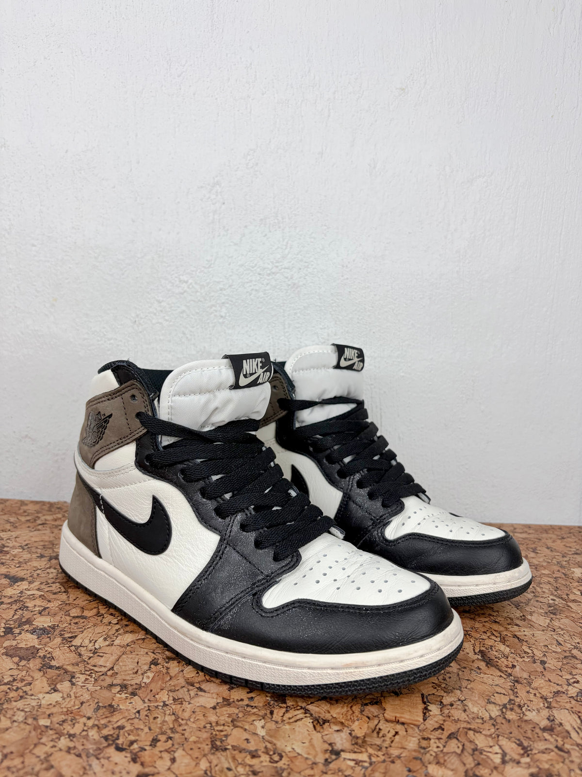 A pair of Air Jordan 1 Retro High OG sneakers in black, white, and brown, displayed on a cork surface against a light gray wall.