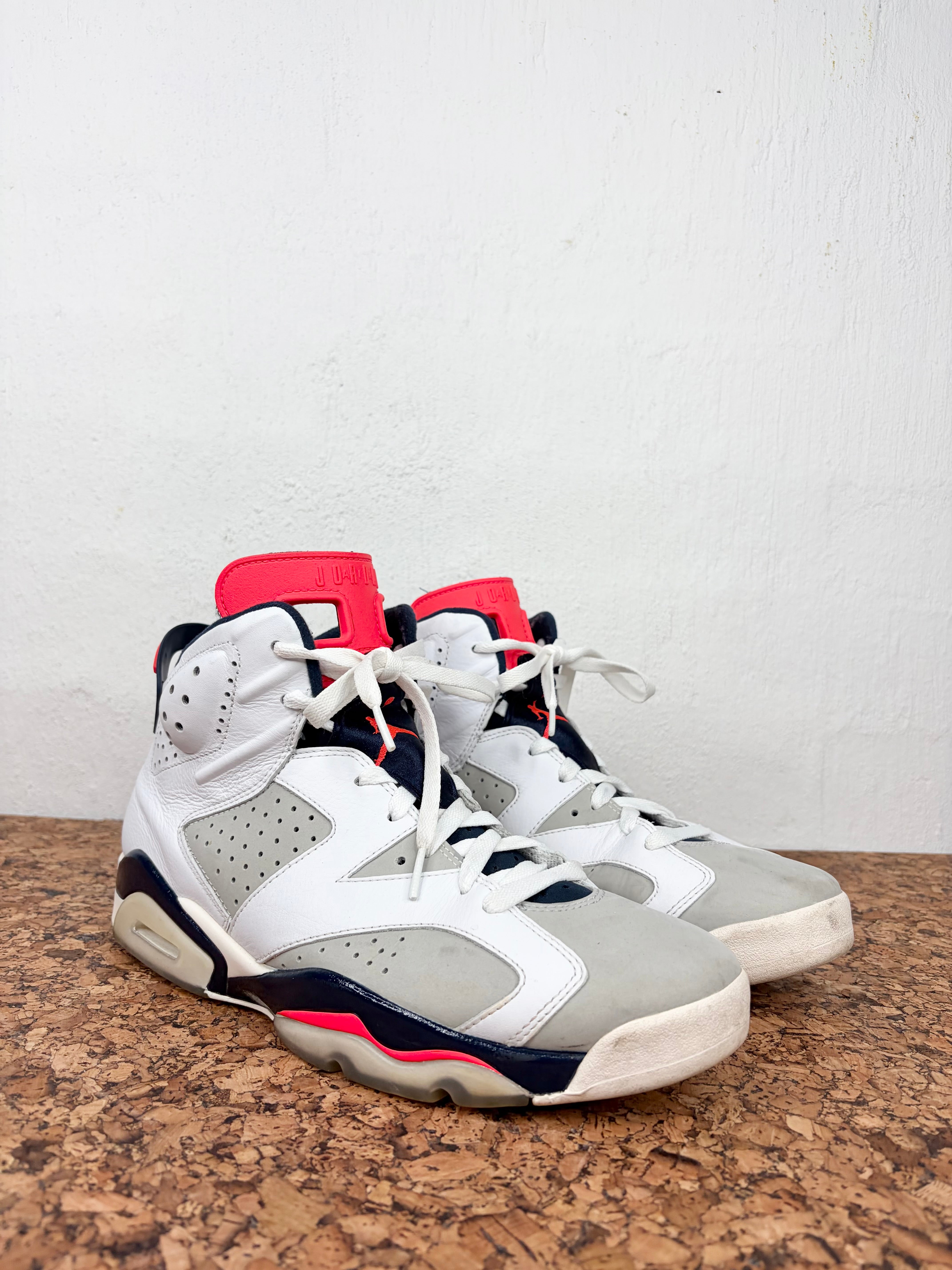 A pair of high-top Air Jordan 6 sneakers in white, gray, and navy blue with bright pink accents, resting on a brown cork mat against a white wall.