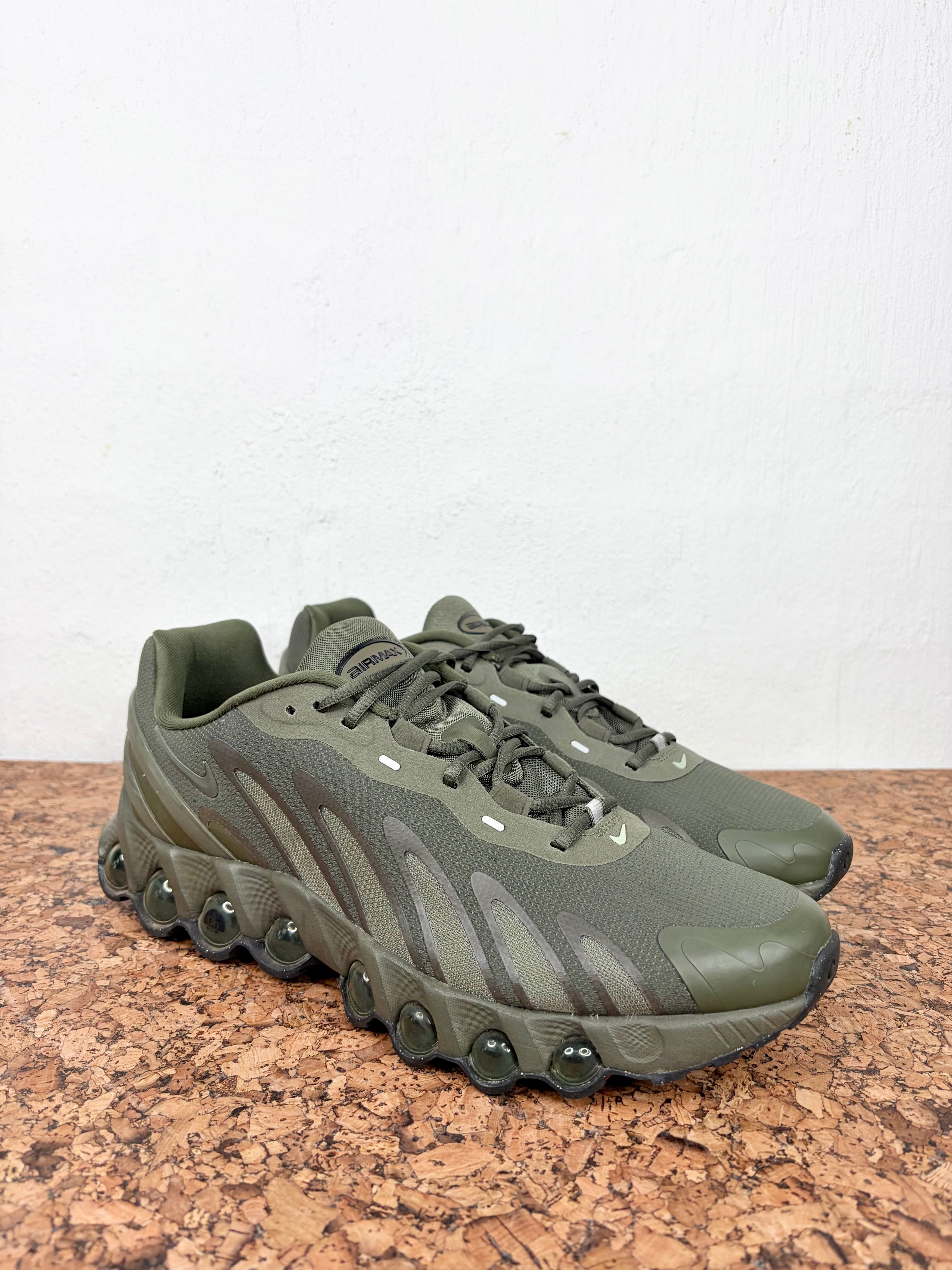 A pair of olive green athletic sneakers with a distinctive wavy sole and bubble-like midsole details, displayed on a cork surface against a white wall.