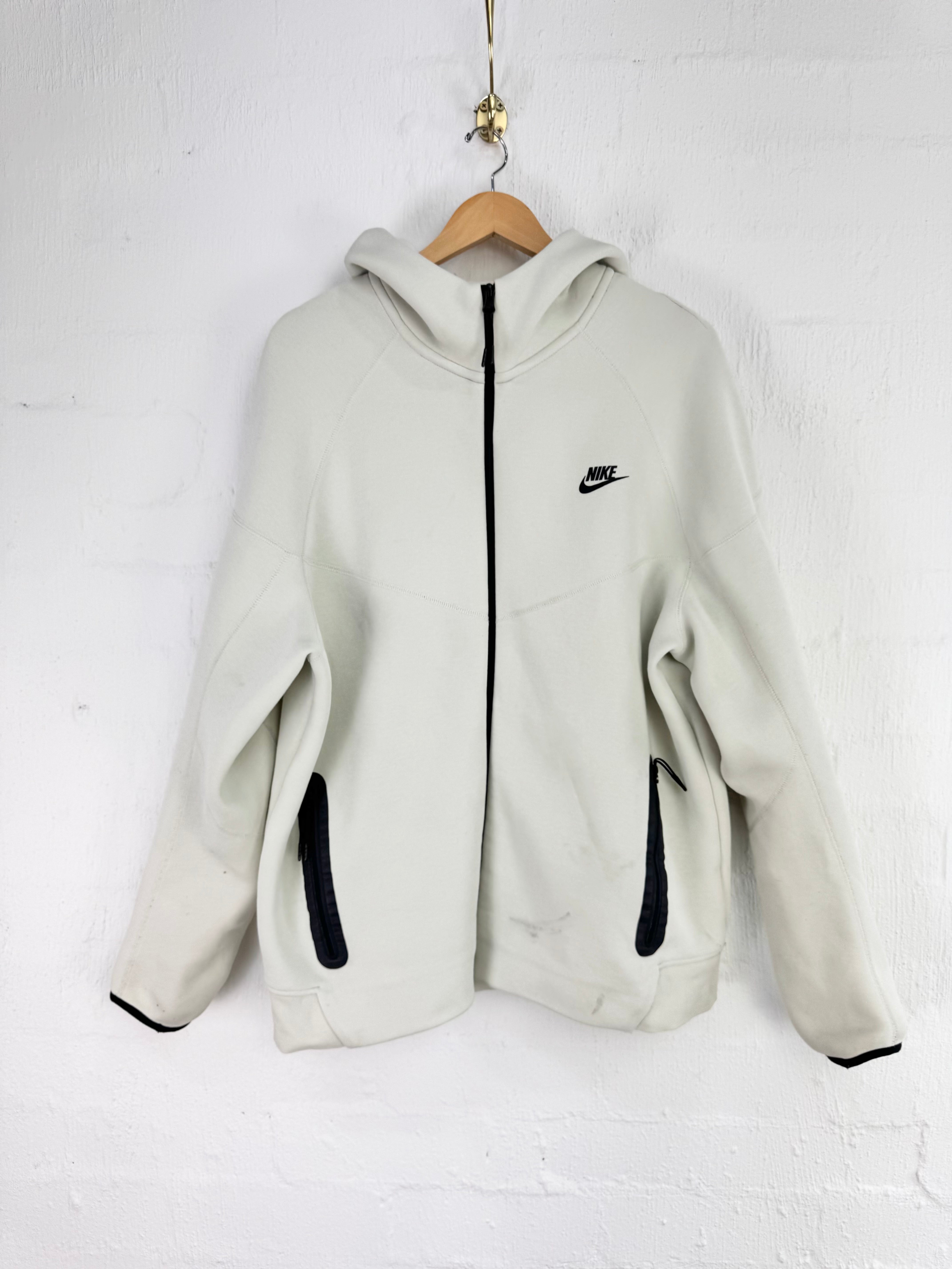 A light beige Nike zip-up hoodie with black accents, hanging on a wooden hanger against a white brick wall.
