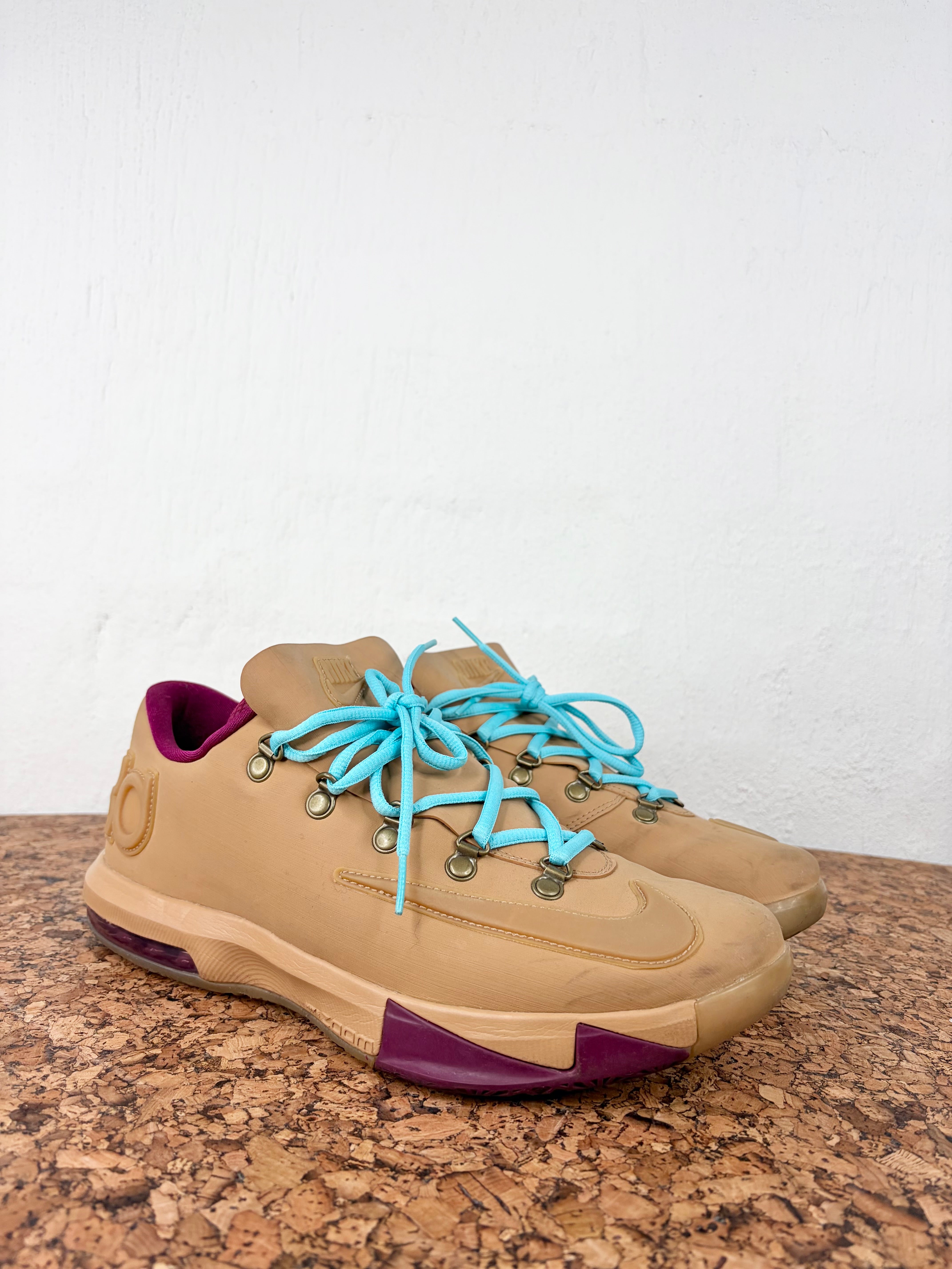 A pair of tan Nike sneakers with bright turquoise laces and purple accents, resting on a cork surface against a white wall.
