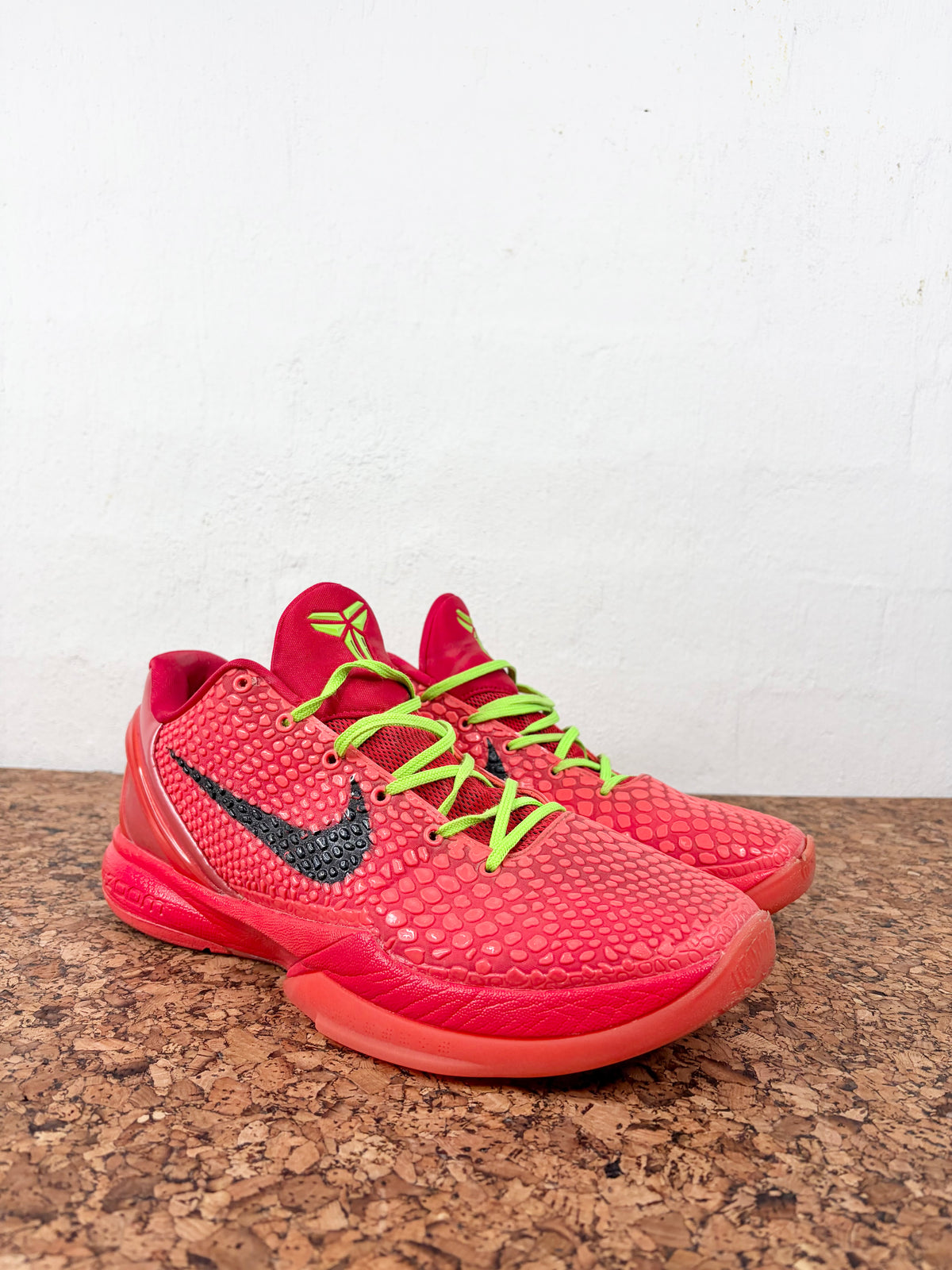 A pair of Nike basketball shoes in a vibrant coral pink and red colorway, featuring a textured knit upper, black Swoosh logo, and bright lime green laces, resting on a brown cork mat against a white wall.