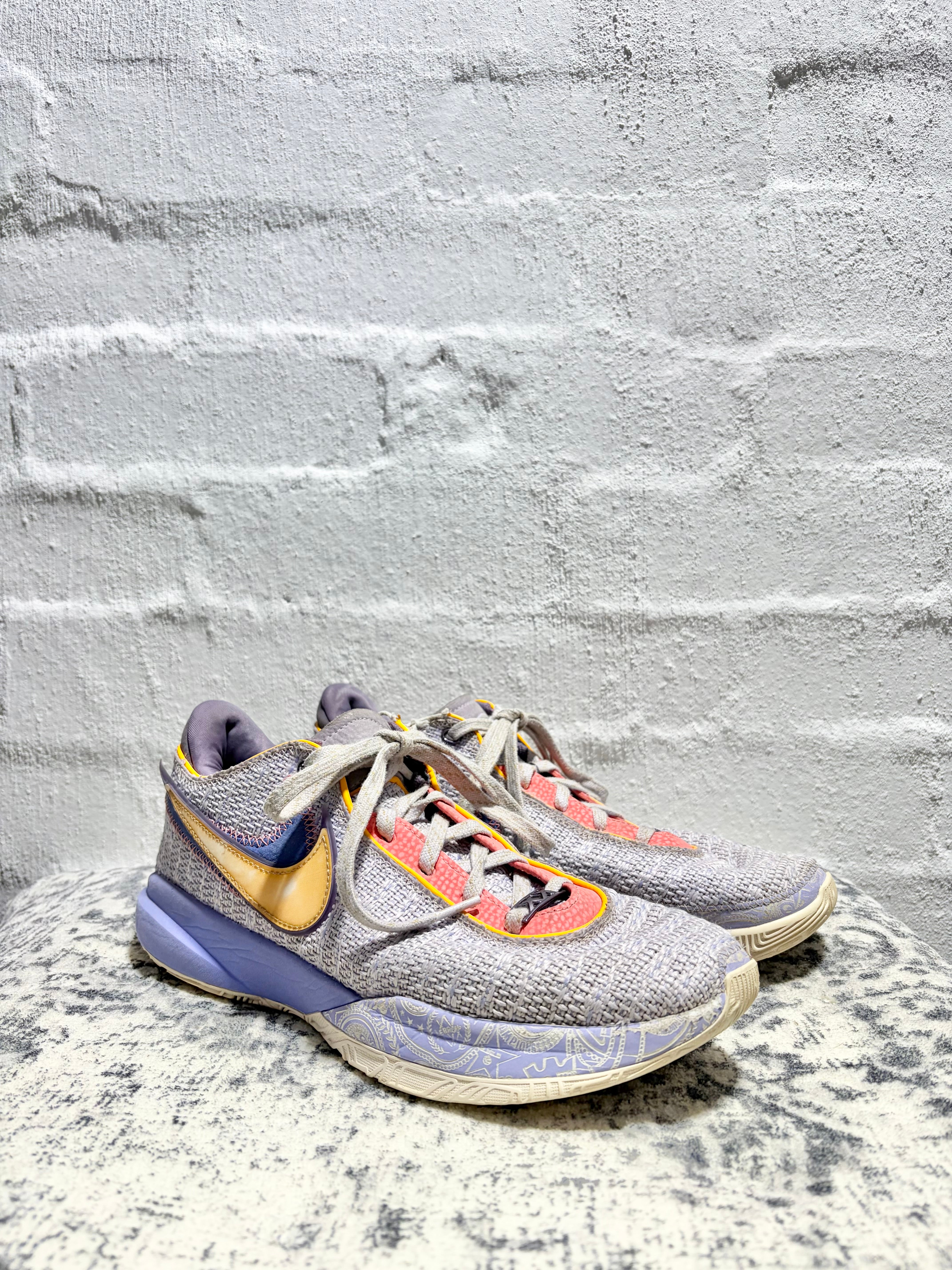 A pair of Nike basketball sneakers in a gray knit upper with purple, gold, and orange accents, resting on a textured stone surface against a white brick wall.
