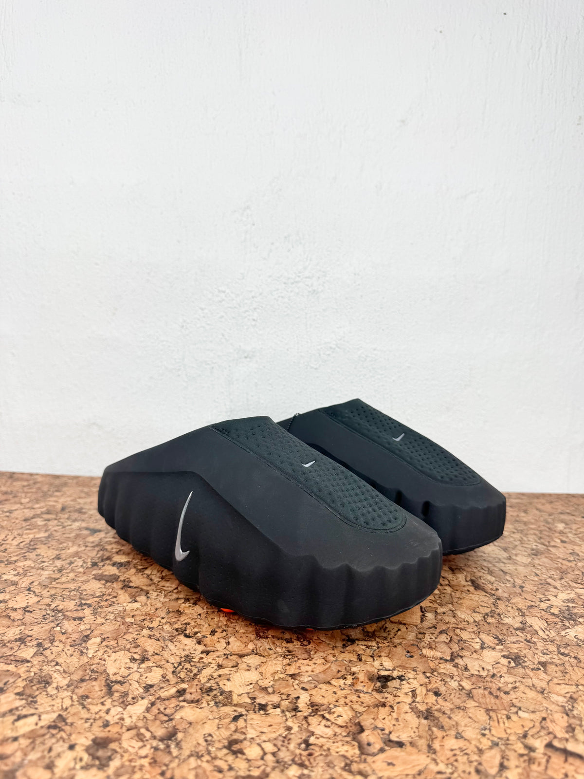 A pair of black Nike slide sandals resting on a brown cork surface against a white wall.