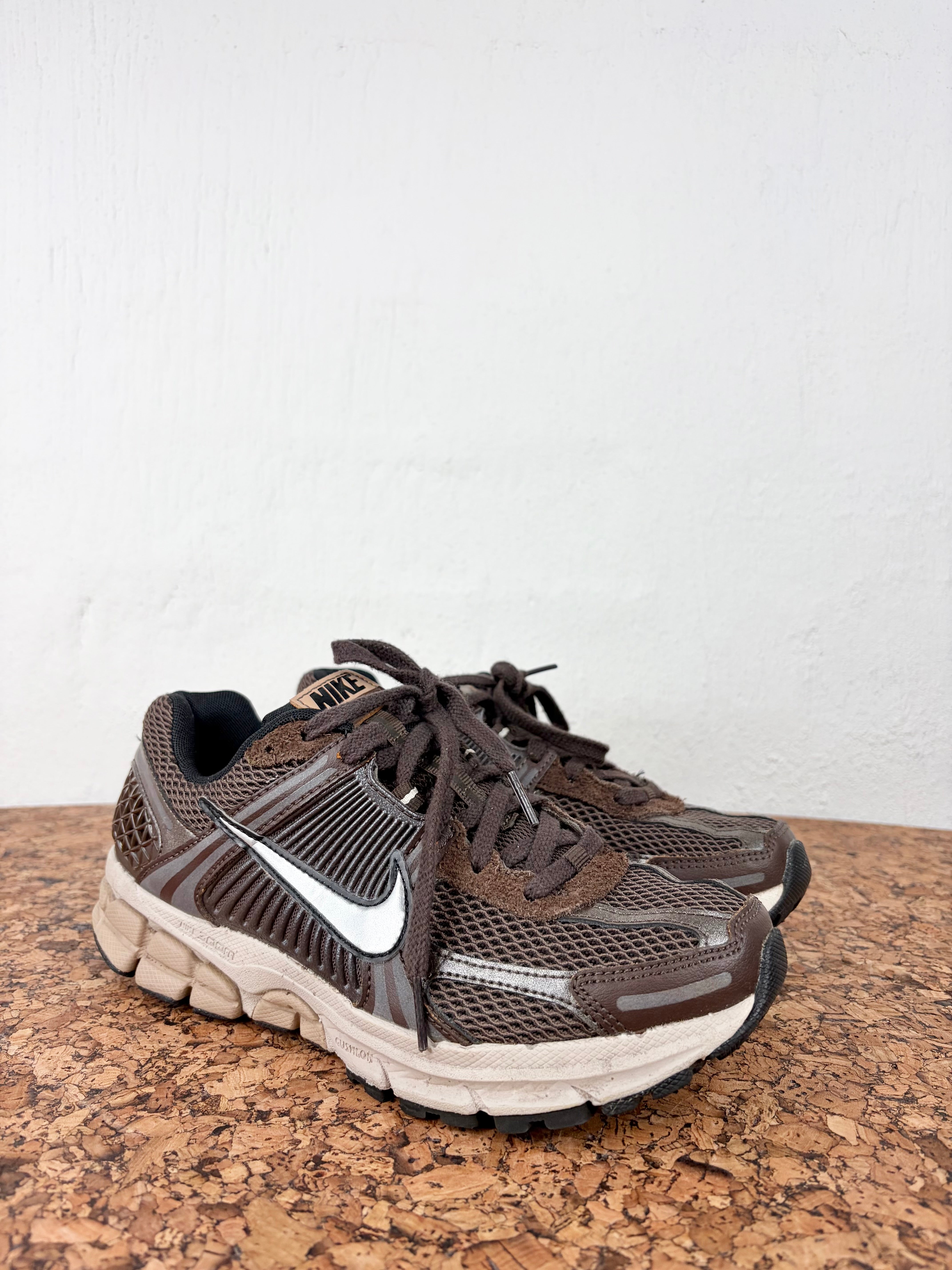 A pair of Nike running shoes in brown and beige with a white Swoosh, resting on a cork surface against a white wall.