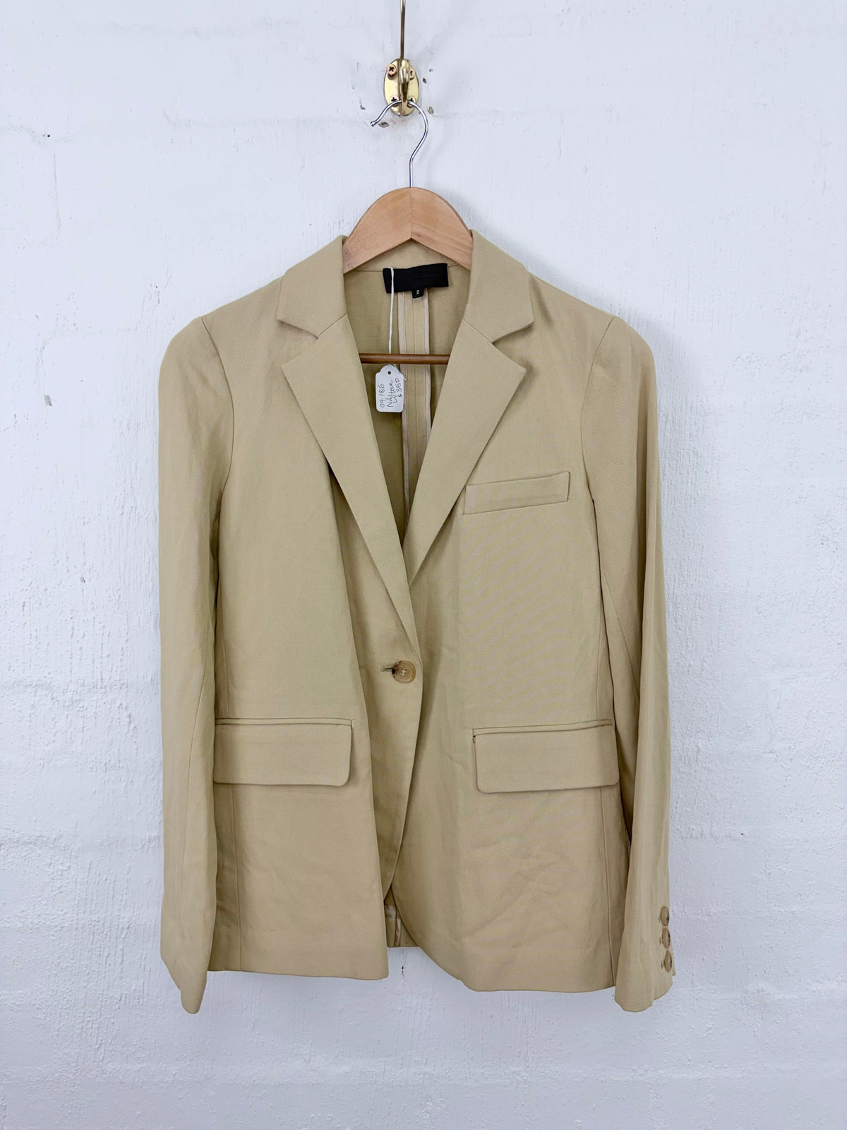 A beige single-breasted blazer jacket with notch lapels and flap pockets, hanging on a wooden hanger against a white painted brick wall.
