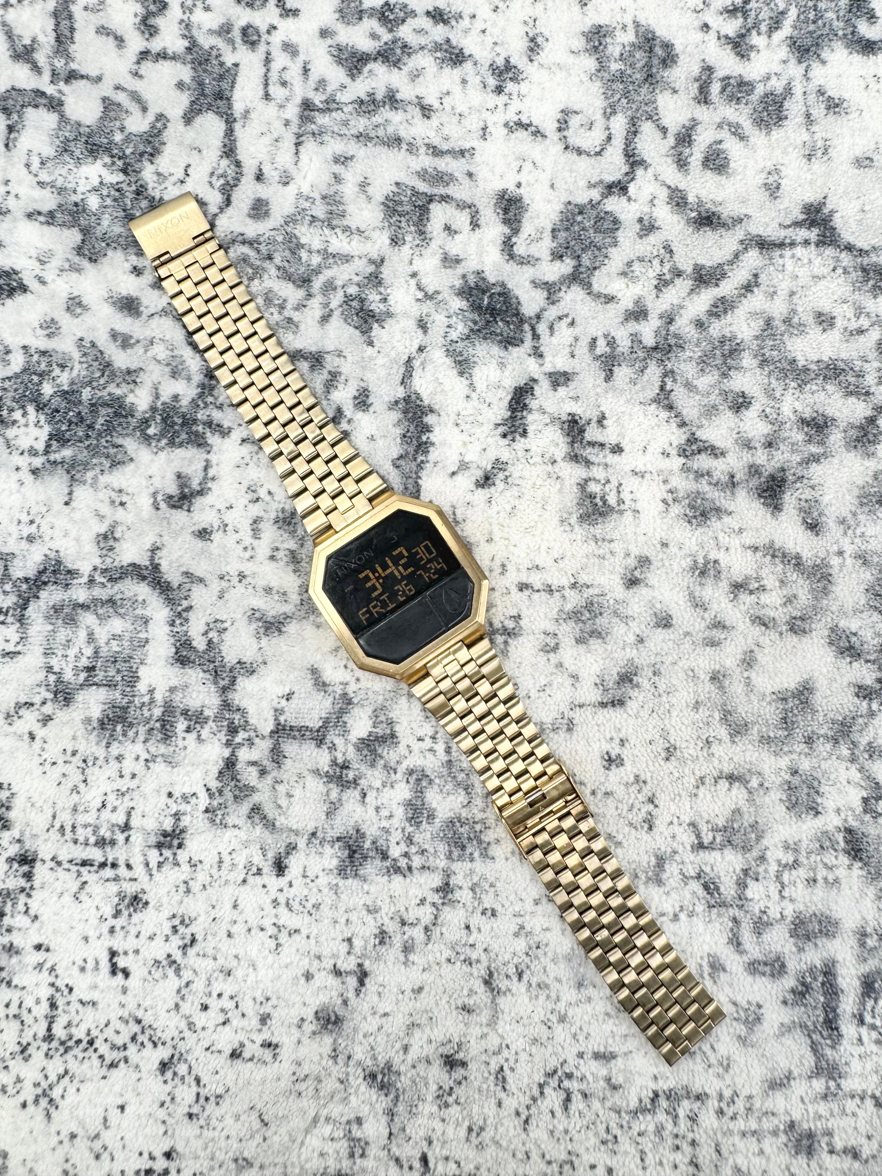 A gold-tone digital watch with an octagonal face and matching metal link bracelet, placed diagonally on a textured gray and white surface.