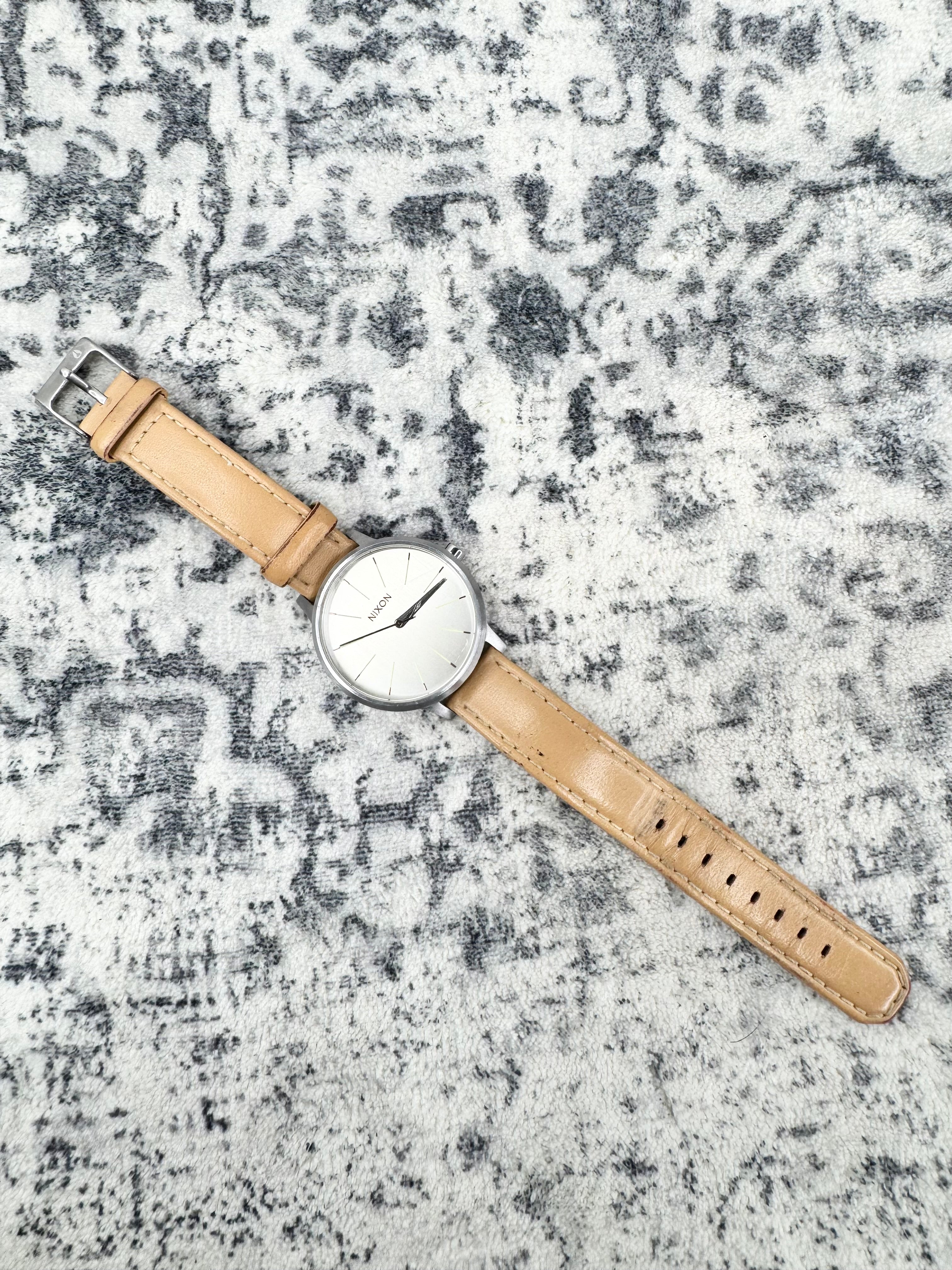 A minimalist wristwatch with a round silver-tone case, white dial, and a light tan leather strap, laid diagonally on a textured gray and white patterned surface.