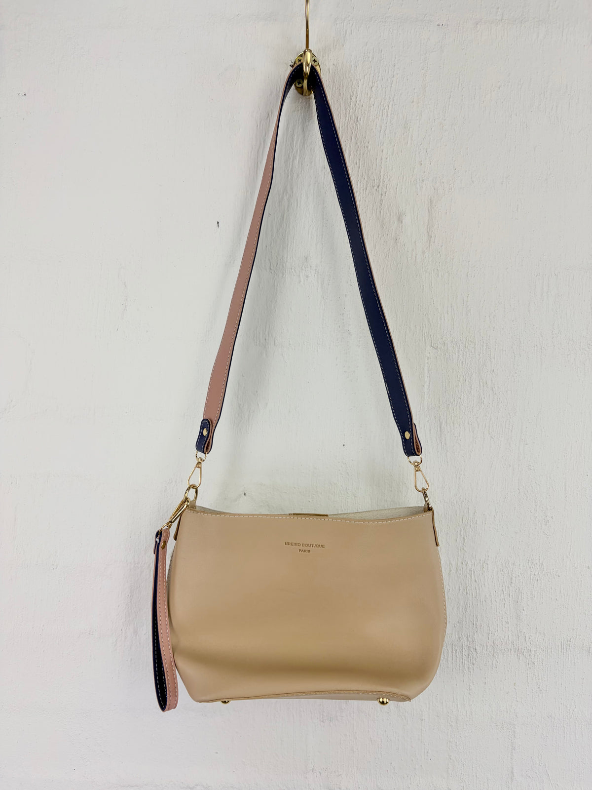 A beige leather shoulder bag with a wide, dual-colored strap featuring beige and navy blue sections, hanging against a white textured wall.