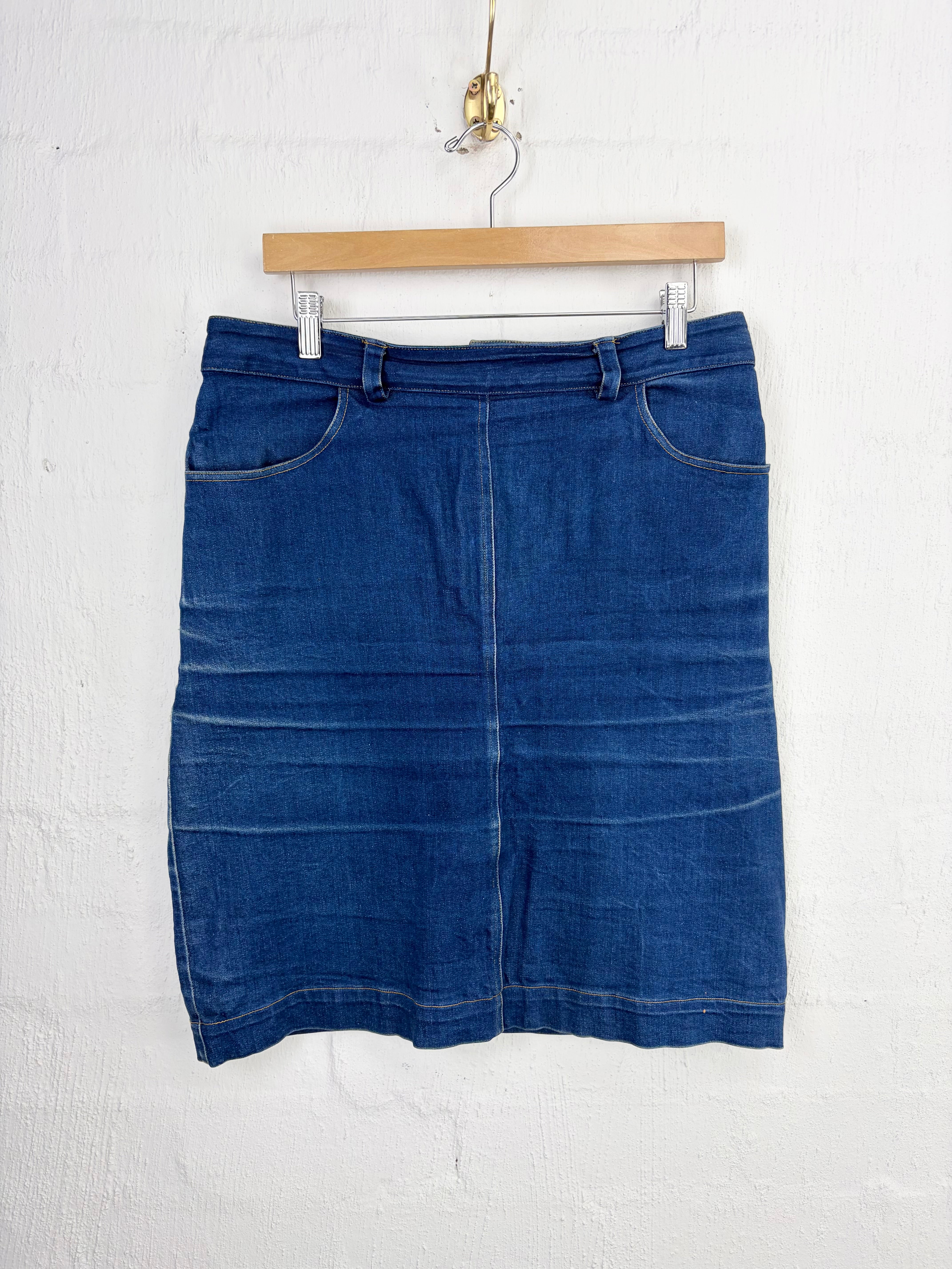 A blue denim skirt with front patch pockets, hanging from a wooden hanger against a white painted brick wall.