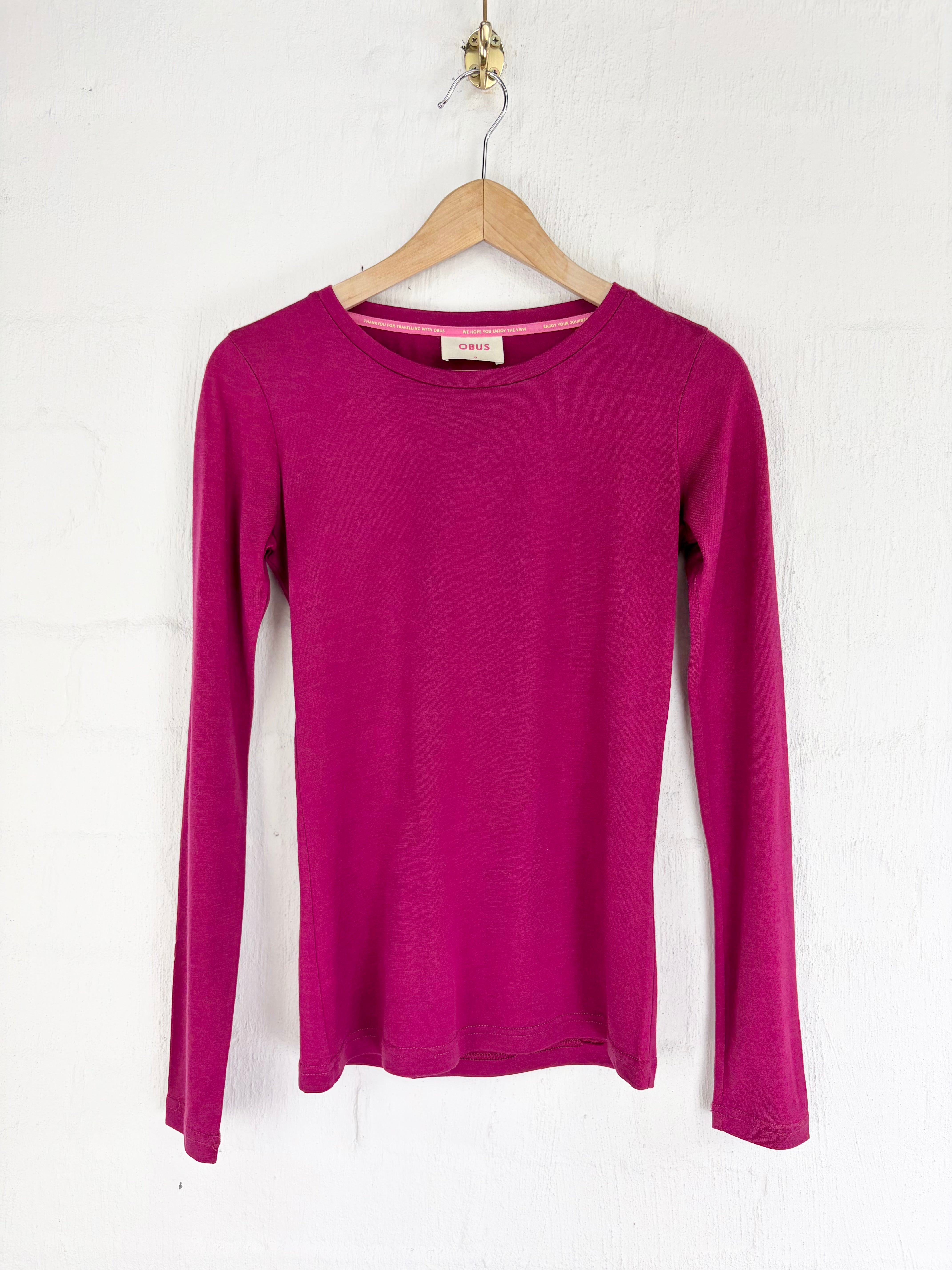 A long-sleeve crewneck knit top in a vibrant magenta pink, hanging on a wooden hanger against a white textured wall.