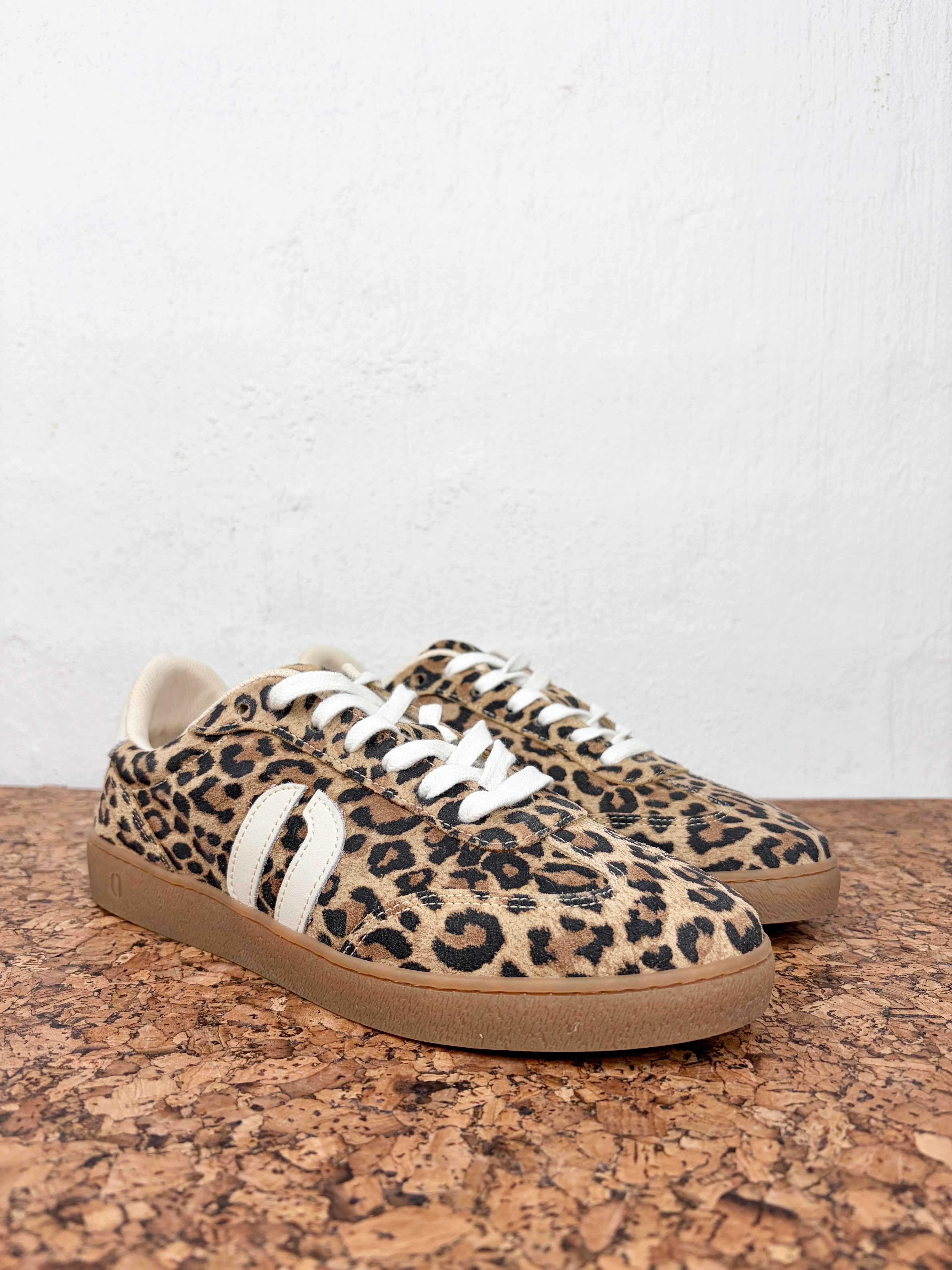 A pair of low-top sneakers with a leopard print pattern in beige, brown, and black, featuring white laces and a white side stripe, resting on a cork surface against a white wall.