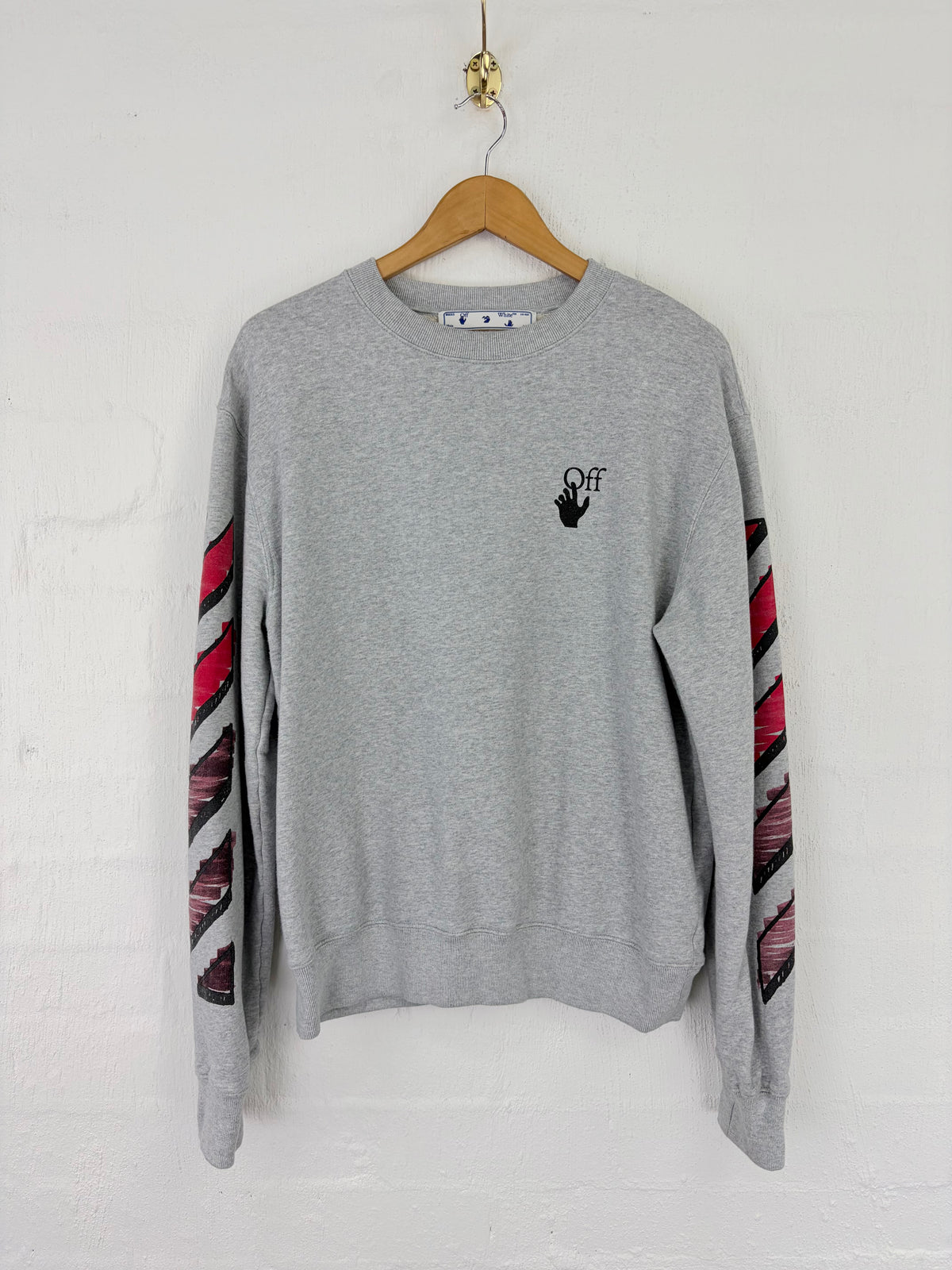 A light gray crewneck sweatshirt from Off-White hangs on a wooden hanger against a white painted brick wall. The sweatshirt features a small black 'OFF' logo with a hand graphic on the left chest and distinctive diagonal striped sleeve patterns in red, black, and white.
