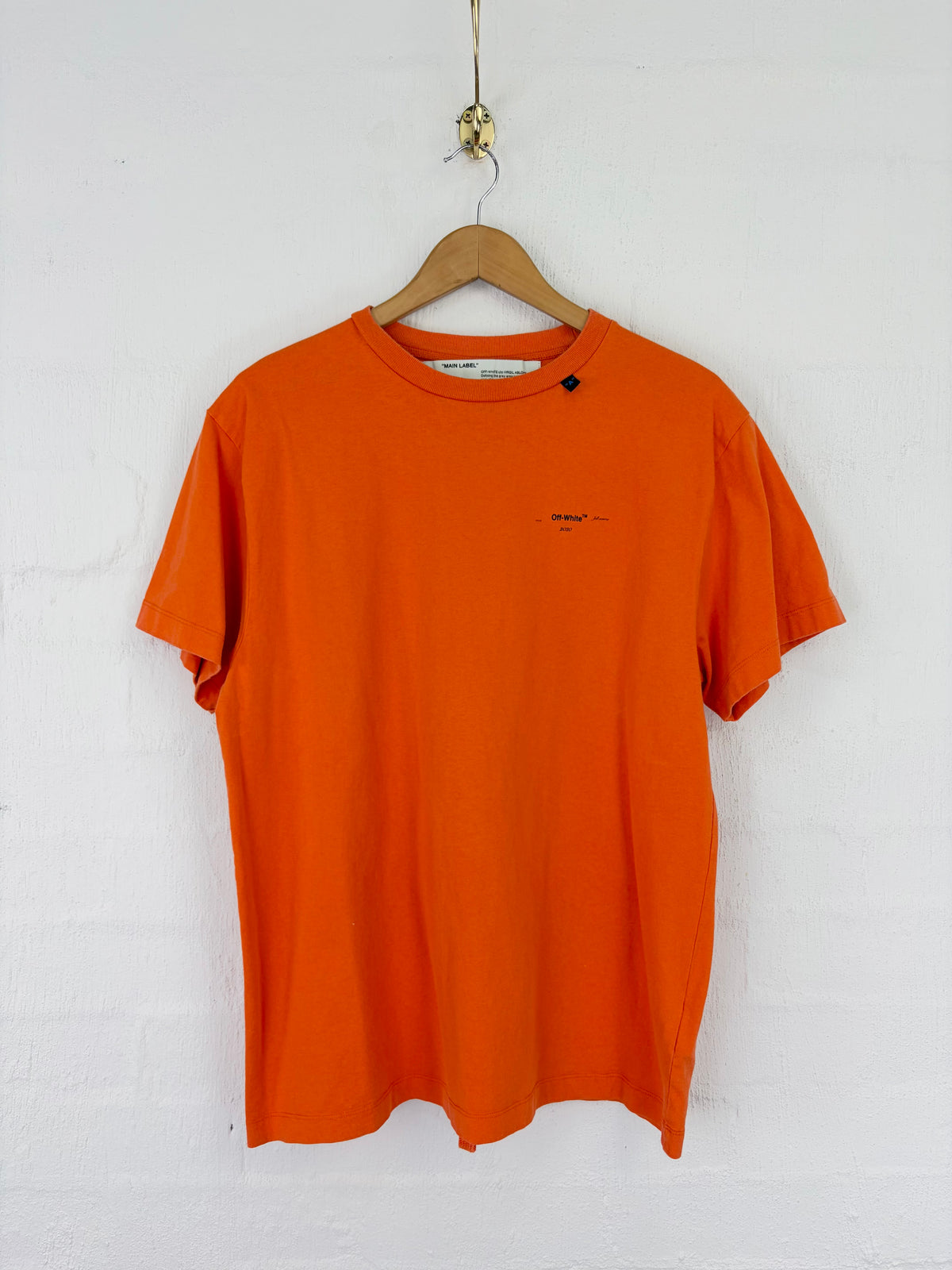 An orange short-sleeve crewneck t-shirt with a small black embroidered logo on the left chest, hanging on a wooden hanger against a white painted brick wall.