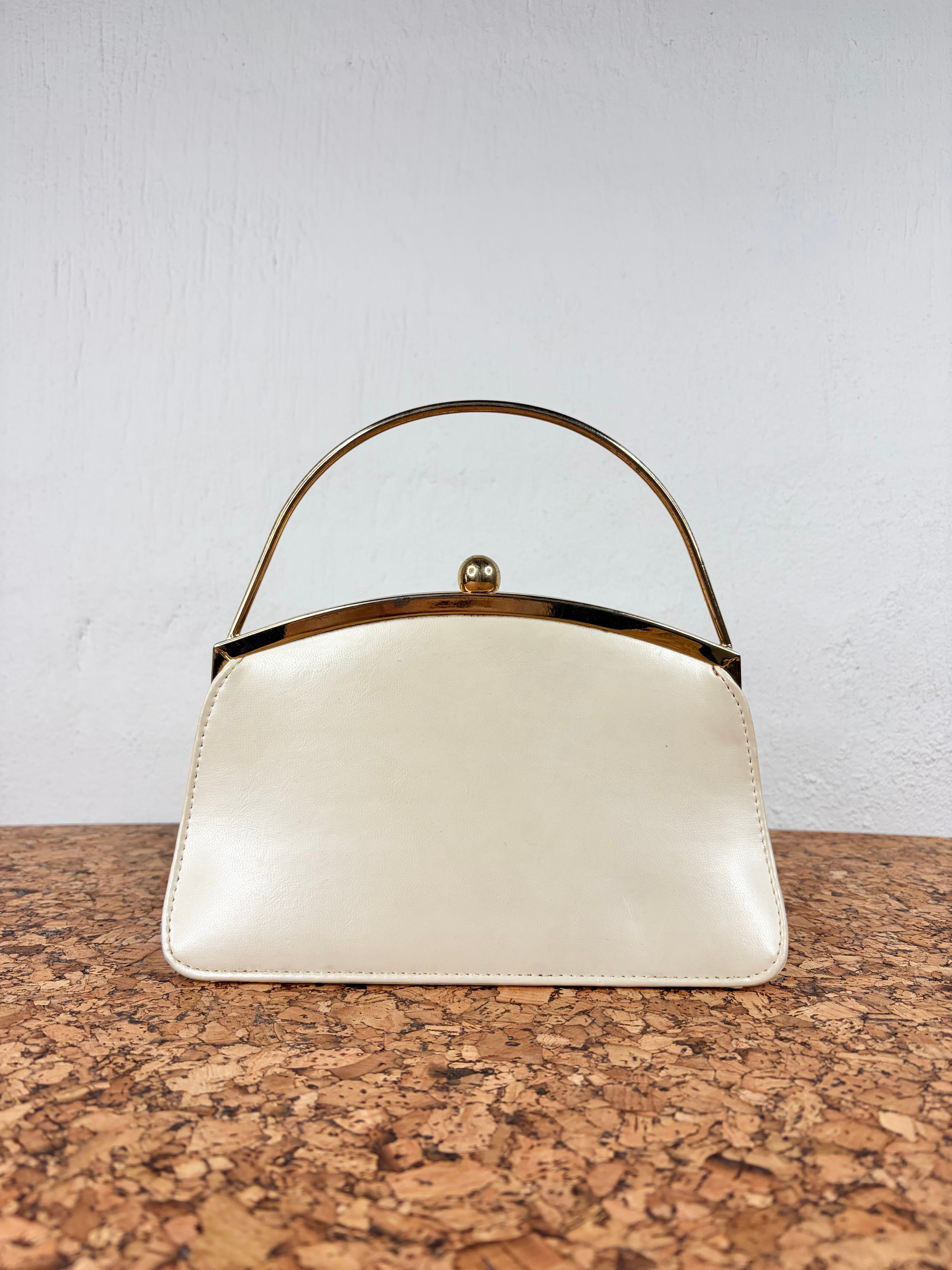 An off-white leather handbag with a gold-tone metal frame and top handle, displayed on a cork surface against a white wall.