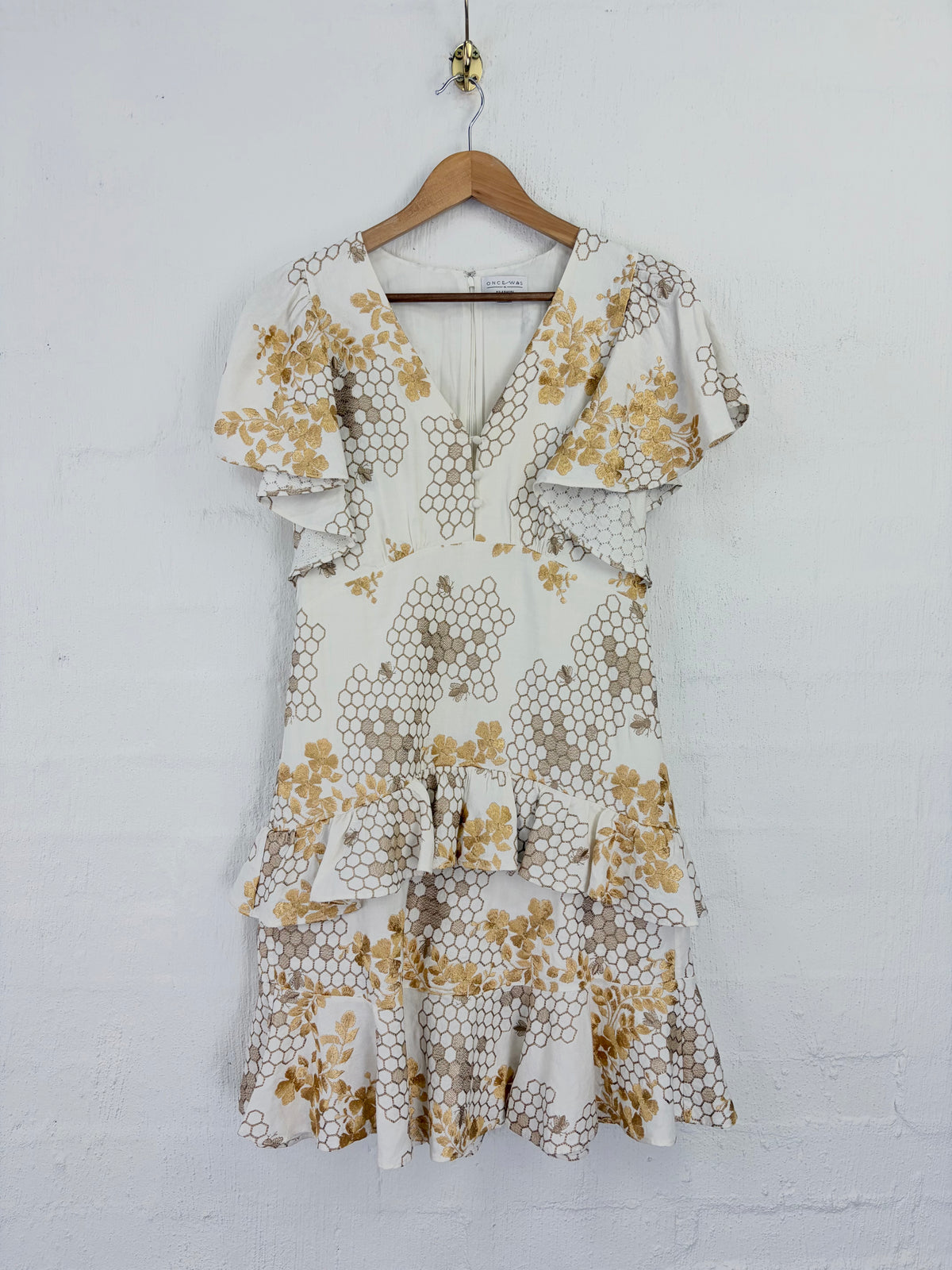 A tiered ruffle mini dress with a honeycomb floral pattern in mustard yellow and taupe on an off-white background, hanging on a wooden hanger against a white painted brick wall.