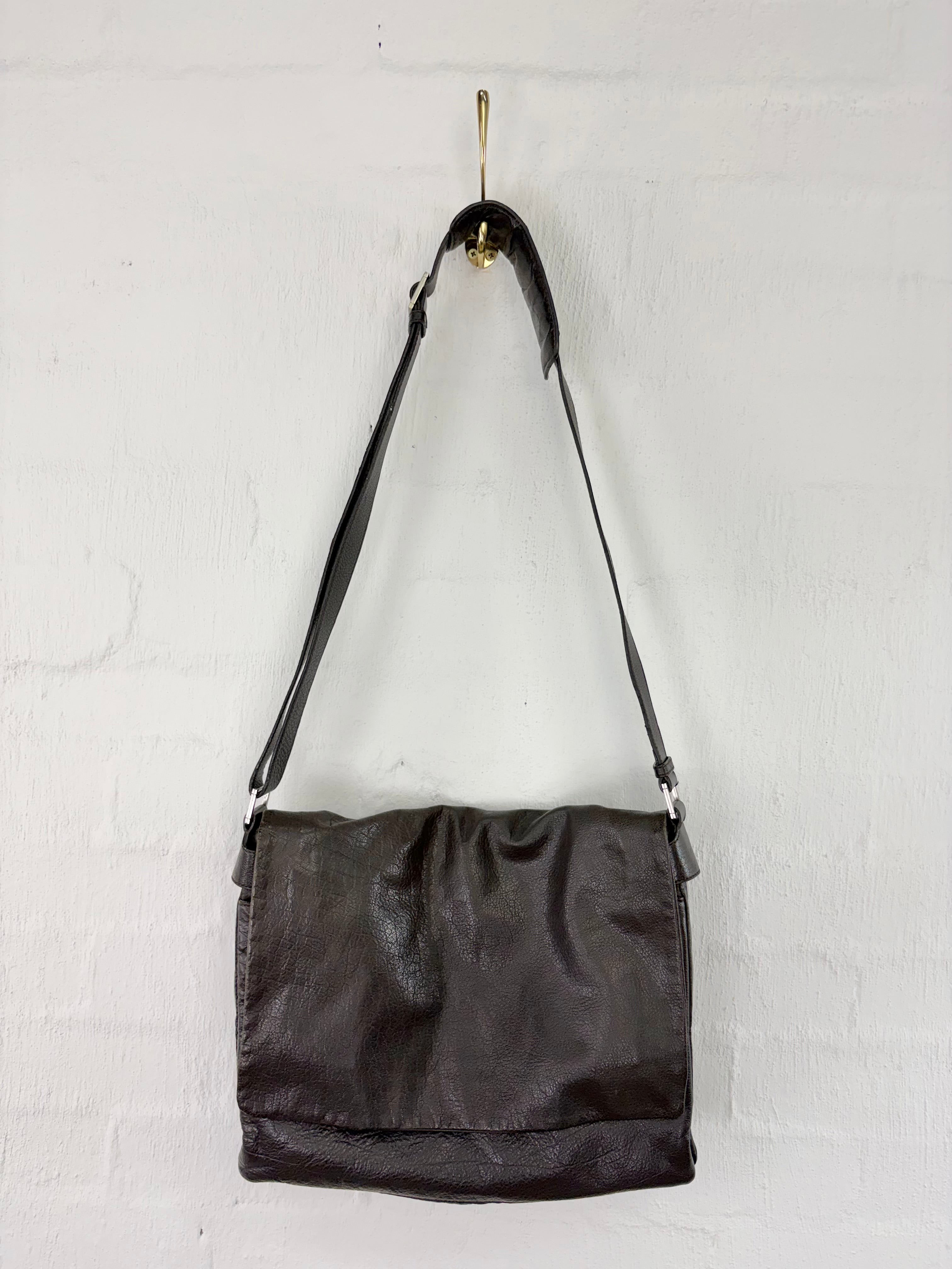 A dark brown leather shoulder bag hanging from a gold hook on a white painted brick wall.