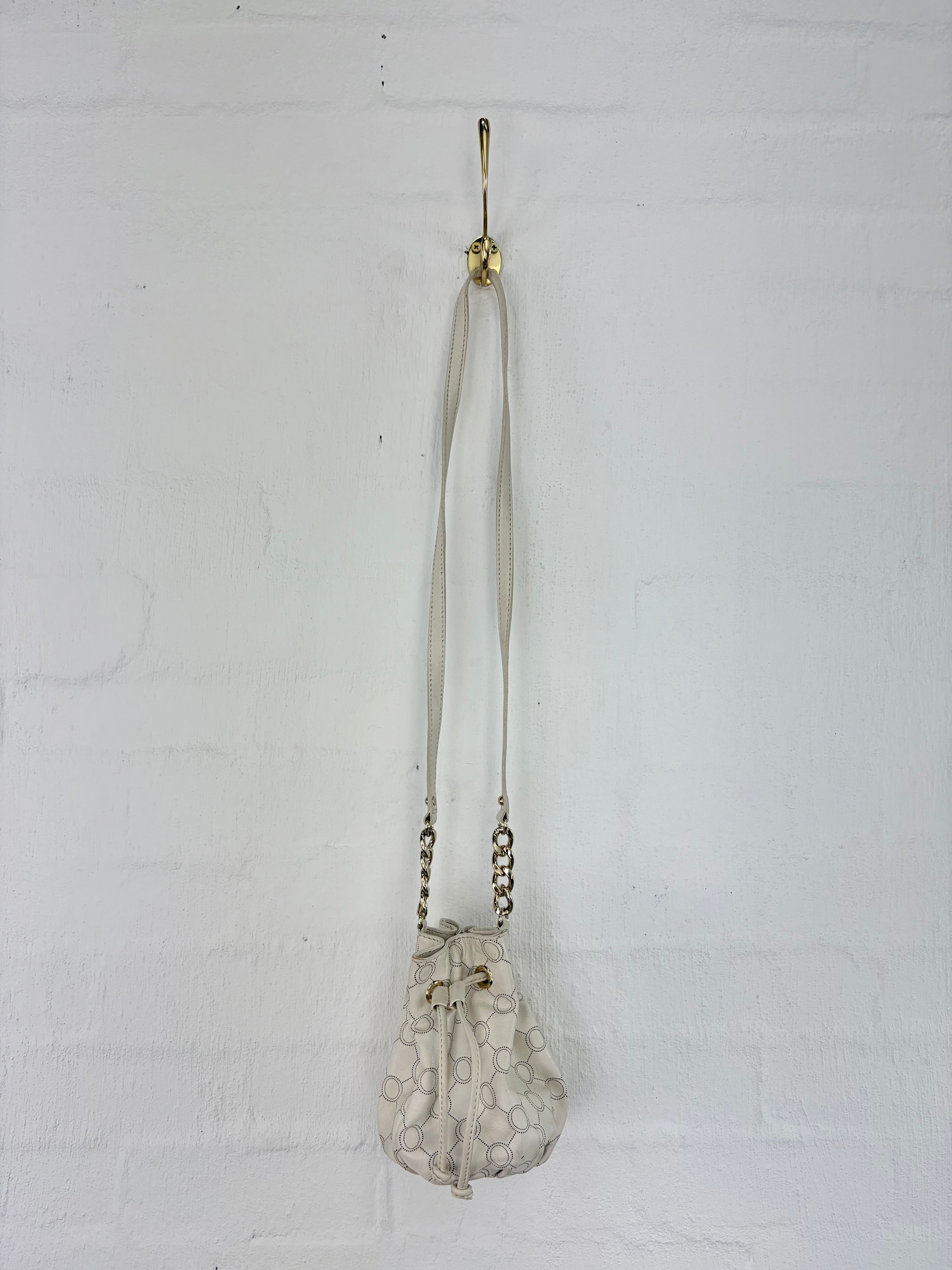 A small beige drawstring pouch bag with an embossed circular pattern and a long strap, hanging from a gold wall hook against a white painted brick wall.