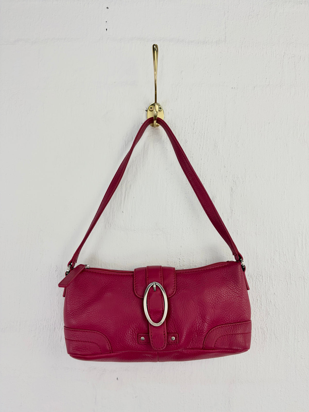 A burgundy pebbled leather shoulder bag with a silver-tone oval ring clasp, hanging from a gold hook against a white textured wall.